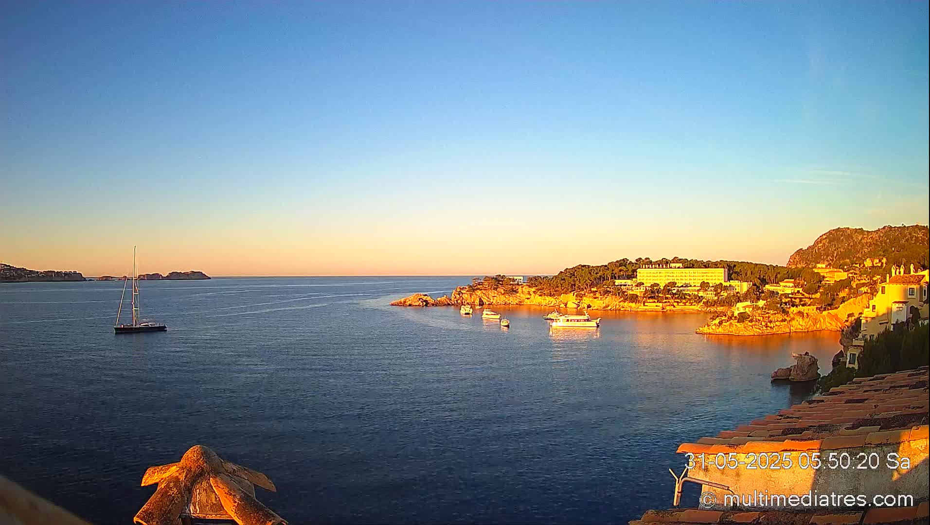 A calm, sunlit ocean cove is seen from a high vantage point, featuring several small boats and a sailboat anchored near a shoreline with buildings and trees.
