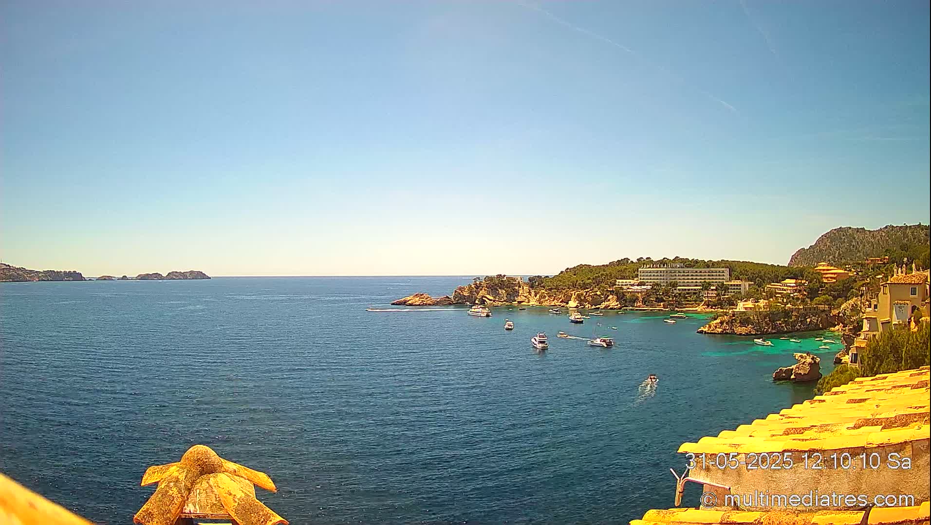 A sunny, clear day overlooking a calm bay with several boats, a hotel, and buildings nestled along a rocky coastline.