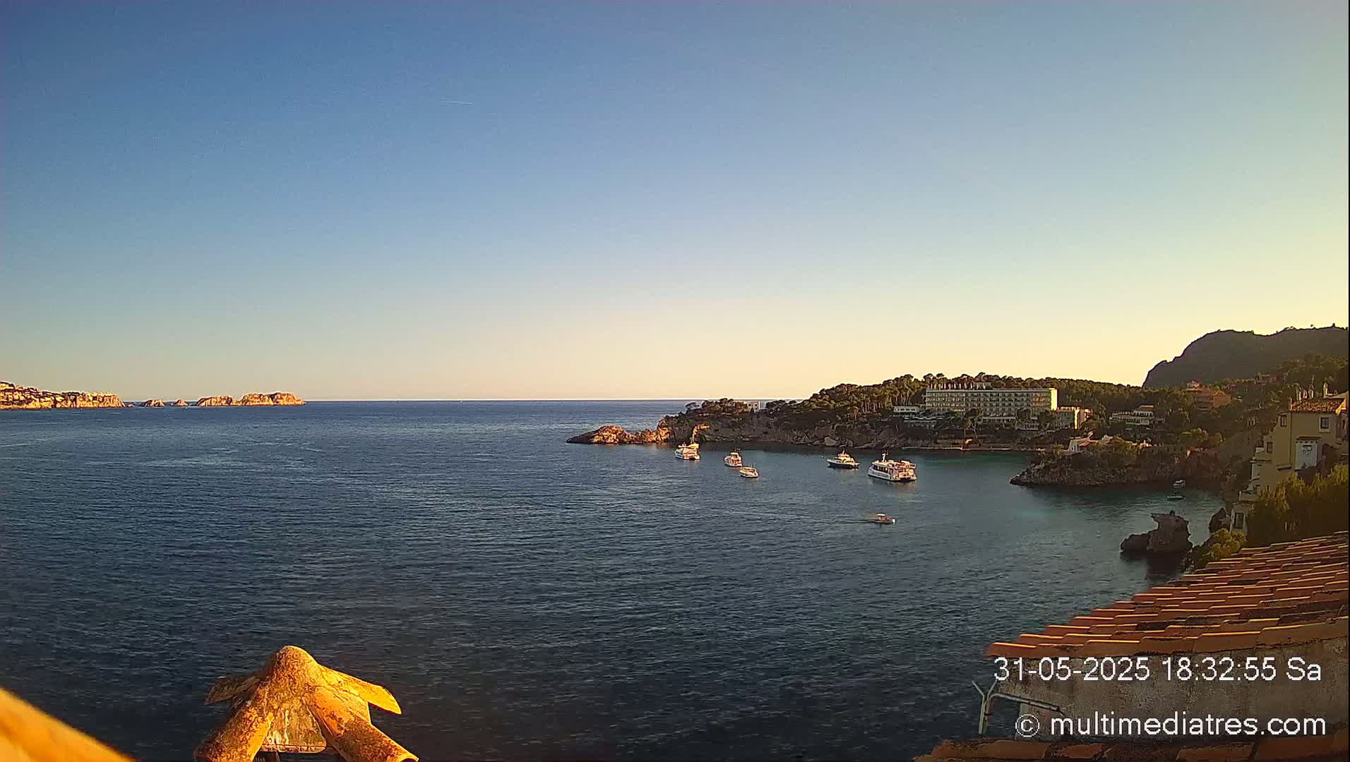 A calm, clear day reveals a coastal scene with several boats anchored in a bay near a building and rocky coastline.