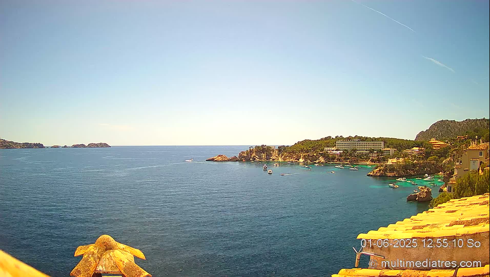 A sunny, clear day shows a calm ocean bay with several small boats, a hotel, and buildings nestled into a rocky, green hillside.