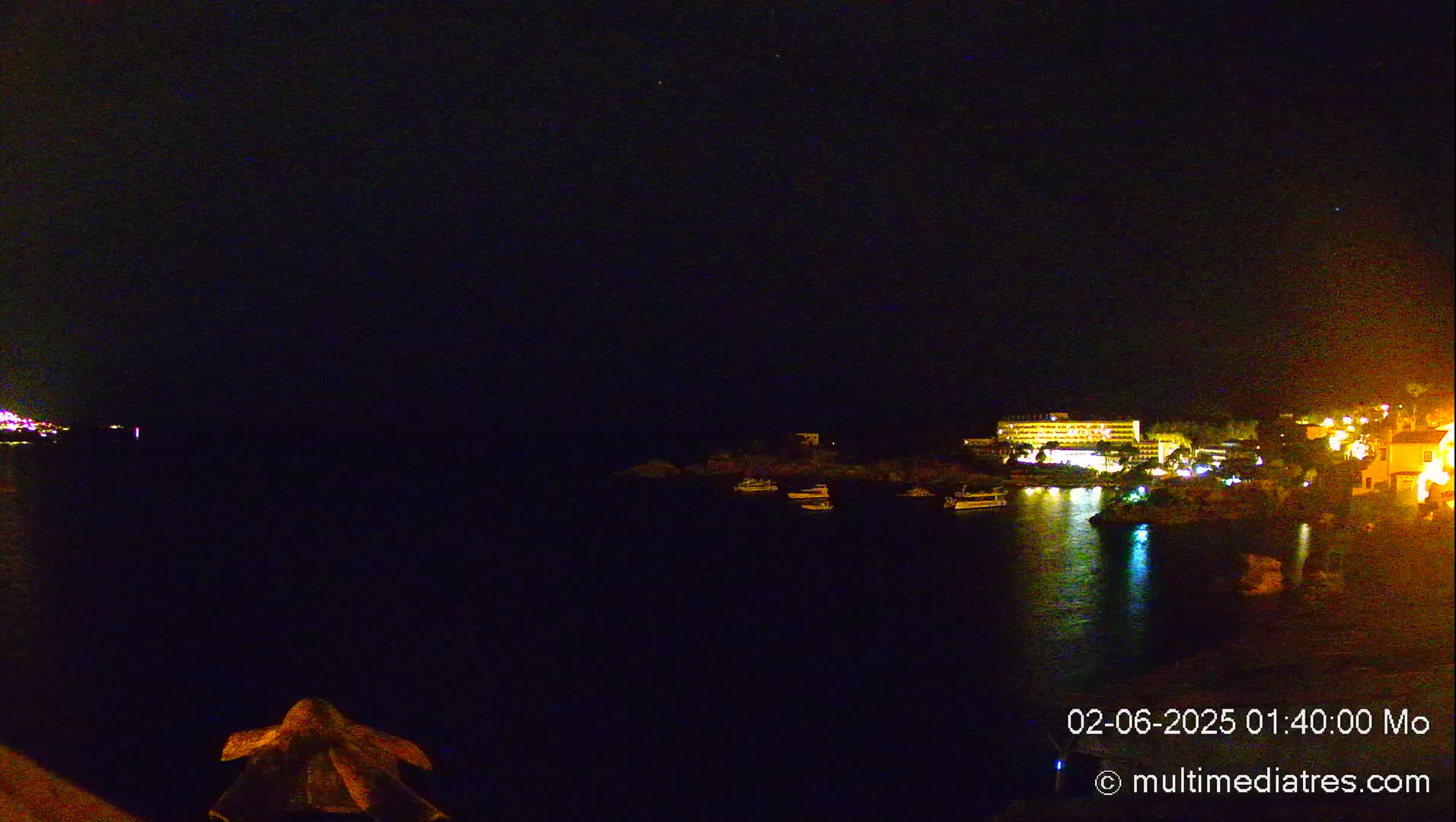 A nighttime, seaside view shows several boats in a dark bay, with illuminated buildings and structures along the shore under a clear, starlit sky.