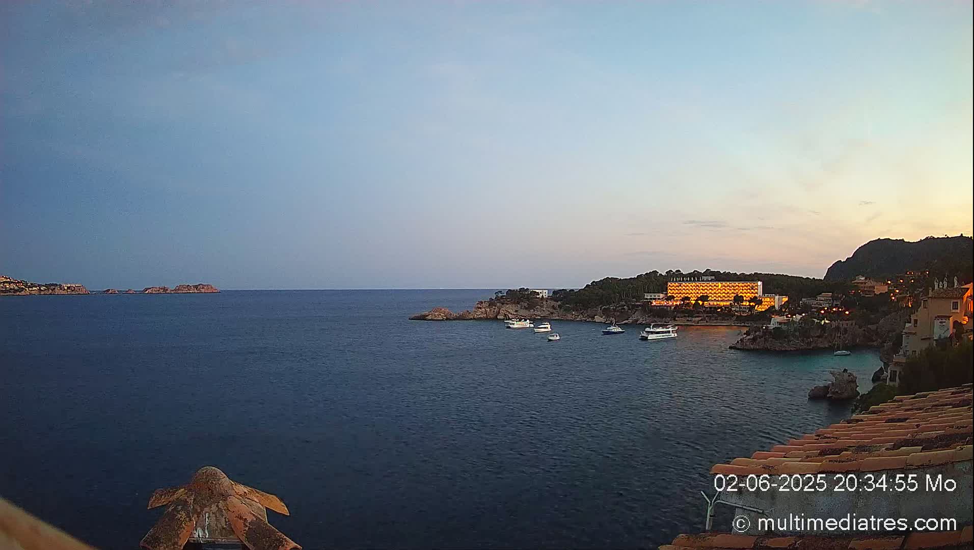 A tranquil coastal scene at dusk shows a calm sea with several boats near a lit-up hotel nestled between rocky shores.