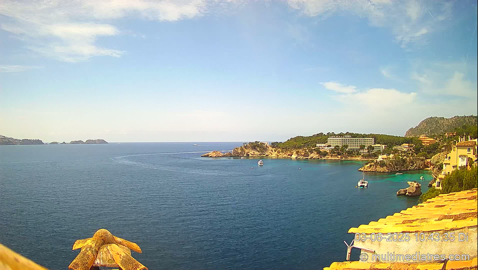 A picturesque coastal scene on a partly sunny day shows a calm blue sea with a few boats, a rocky shoreline with buildings and trees, and small islands in the distance.
