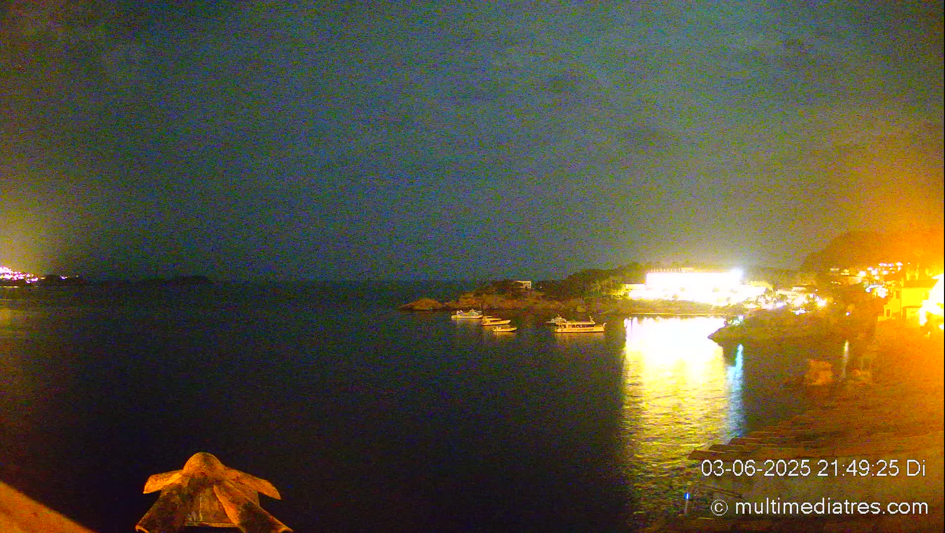 A nighttime view of a calm body of water with several boats and a brightly lit shoreline.