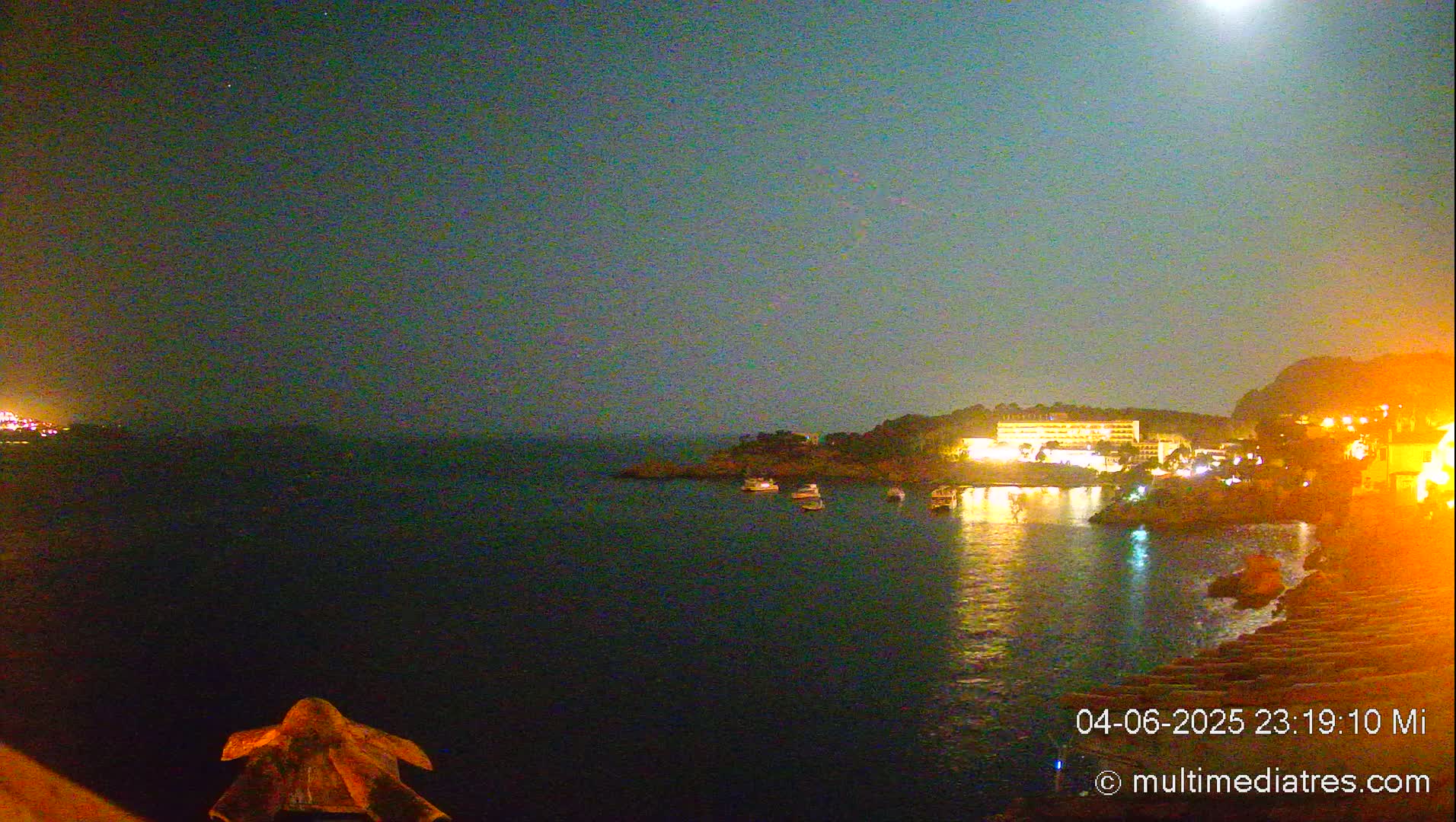 A nighttime seaside view shows a calm body of water reflecting the lights of buildings on the shore under a clear, moonlit sky.