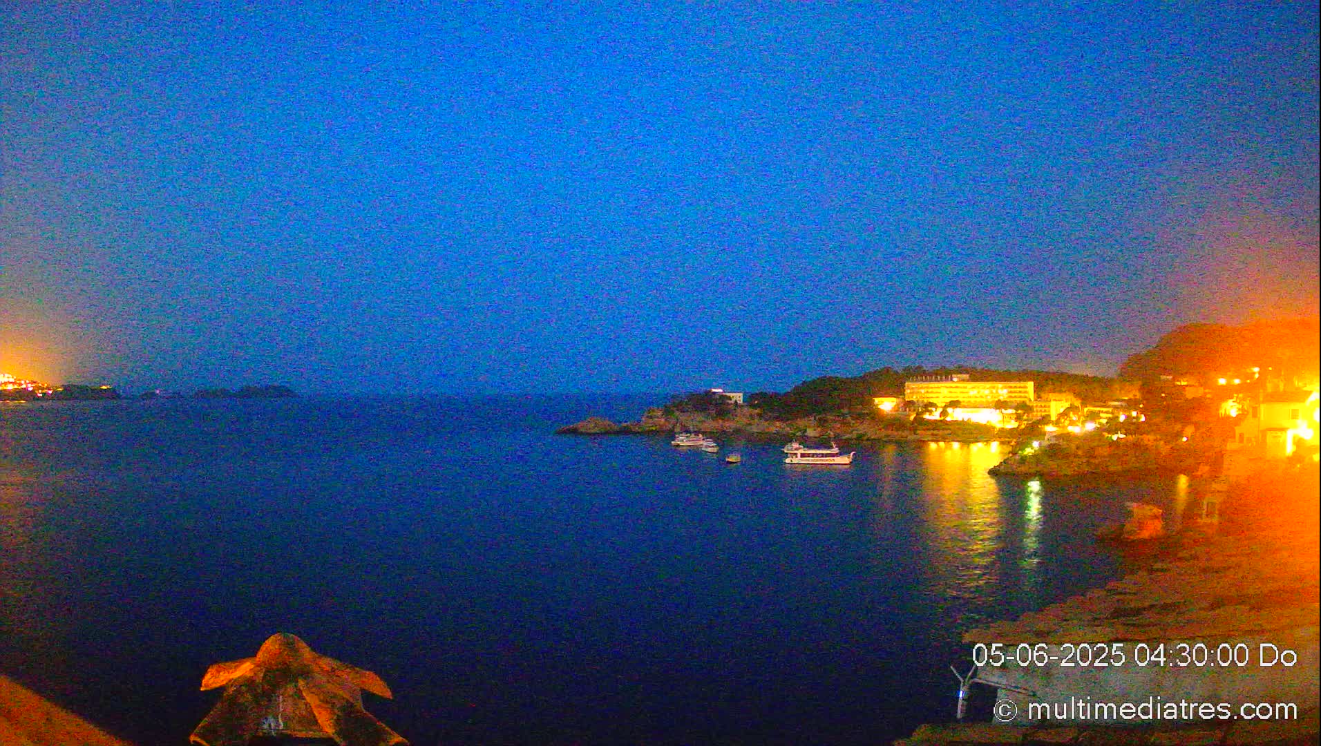 A dark-blue, twilight sky overlooks a calm body of water at night, where several boats are moored near a brightly lit coastal town.