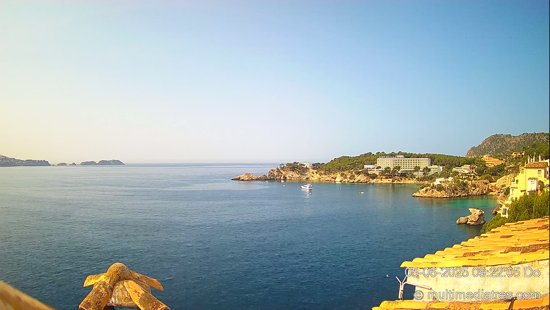 A calm, clear day shows a coastal scene with a body of water, rocky shores, buildings, and some boats.
