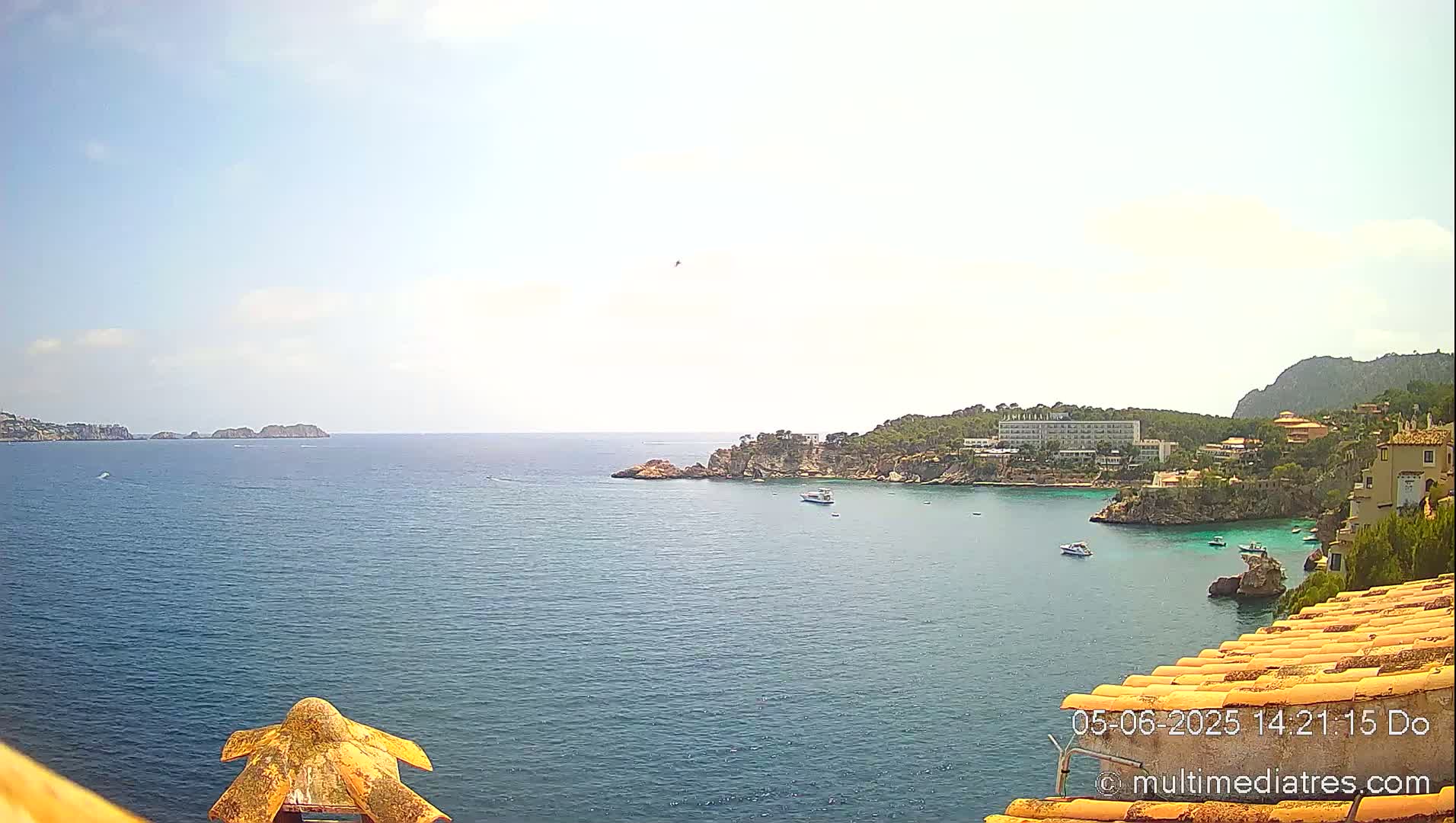A sunny day overlooks a calm, blue ocean bay with small boats, rocky shorelines, and buildings nestled into the hillside.