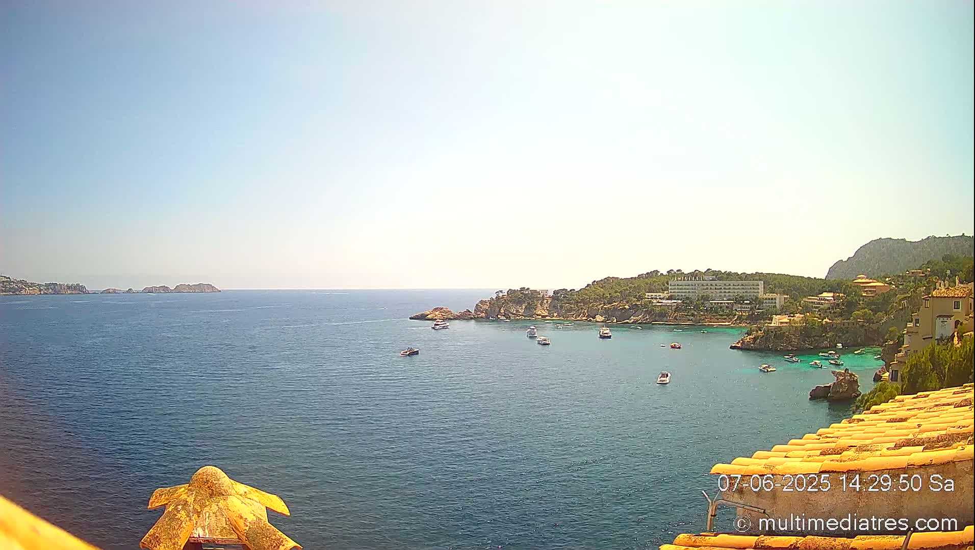 A sunny, clear day overlooks a calm bay with several boats and a coastline featuring buildings and green vegetation.
