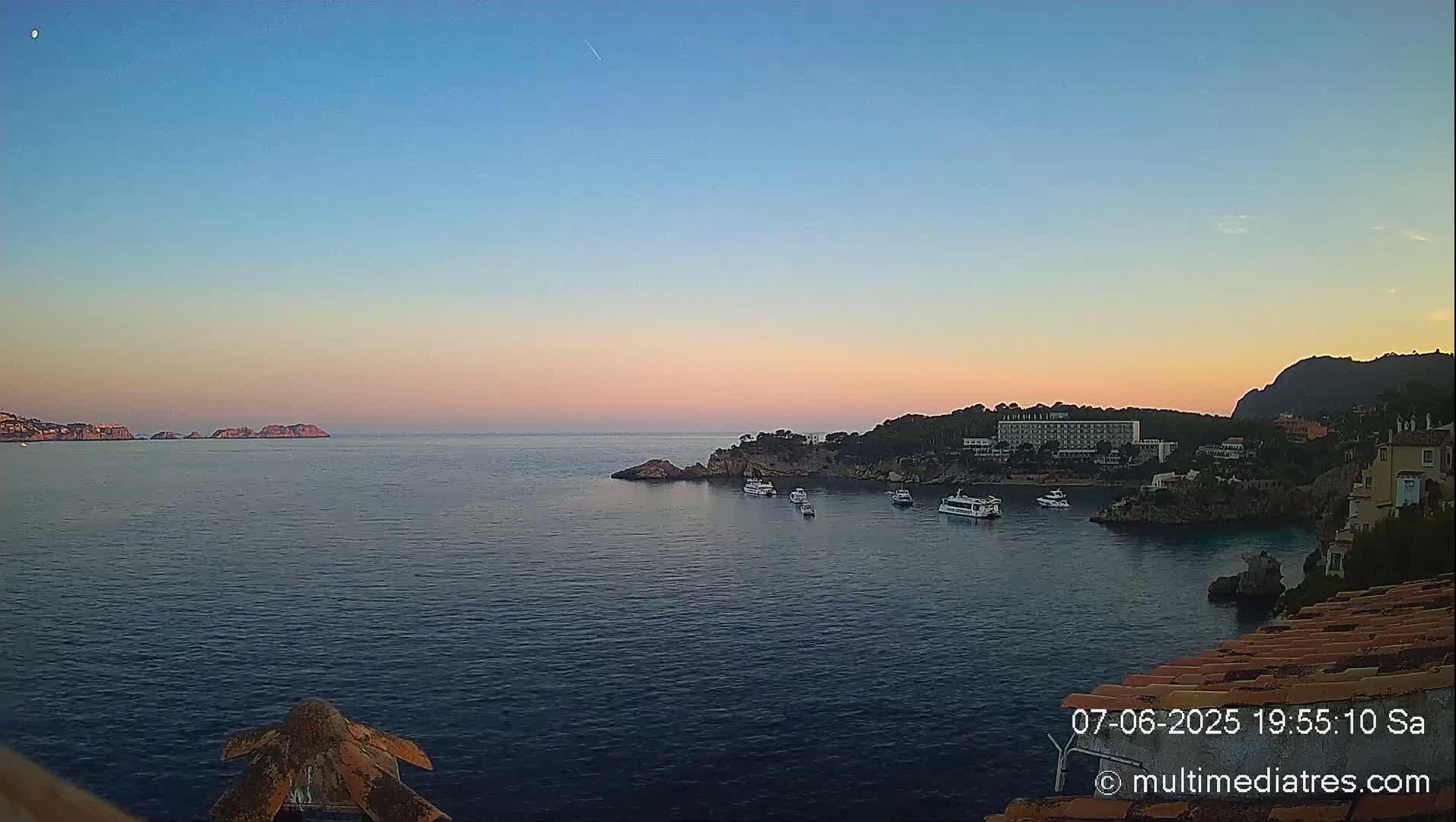 A tranquil coastal scene at sunset shows a calm sea with several boats near a rocky shoreline and a large hotel, under a clear, pale sky.
