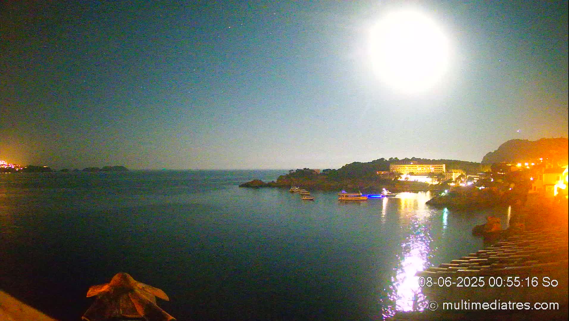 A brightly lit full moon shines over a dark body of water with several boats and a coastline featuring buildings at night.