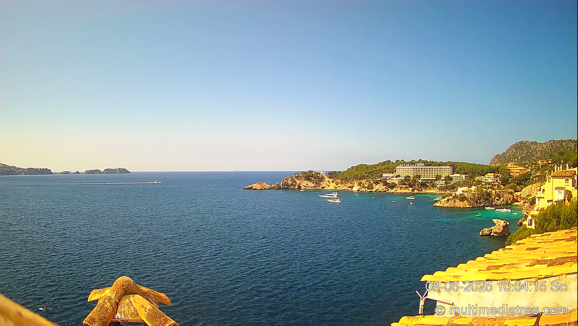 A tranquil blue ocean cove is seen from a high vantage point on a sunny day, with boats scattered in the calm water and buildings nestled along a rocky coastline.