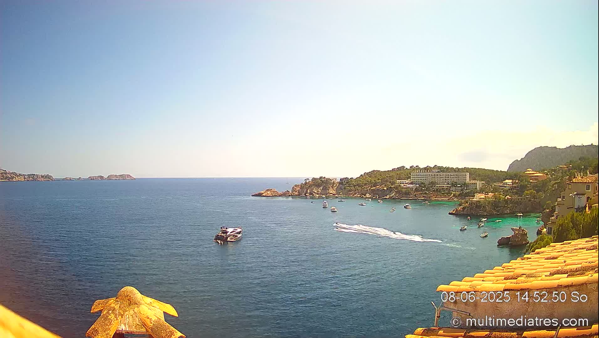A sunny day reveals a calm, blue bay with several boats, a rocky coastline with buildings, and distant islands under a clear sky.