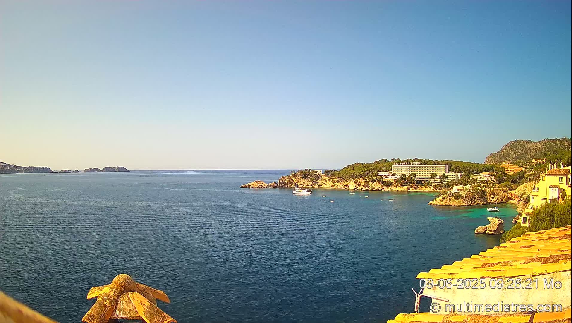 A clear, sunny day reveals a tranquil bay with a calm sea, a small boat, and buildings nestled along a rocky coastline.