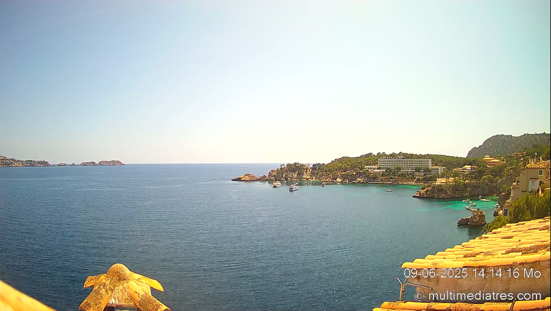 A sunny coastal scene shows a calm blue sea with a small bay containing boats and buildings nestled among rocky hillsides.