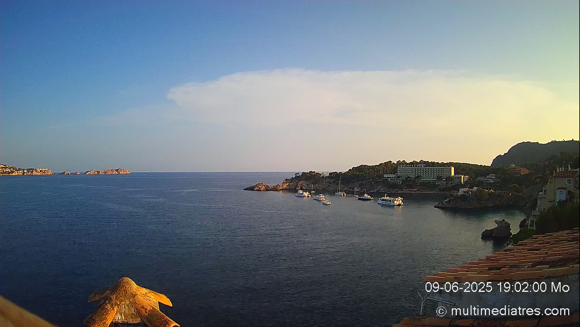 A calm, clear blue sea with several boats near a coastline featuring a hotel and other buildings under a mostly clear sky with a single, thin cloud.