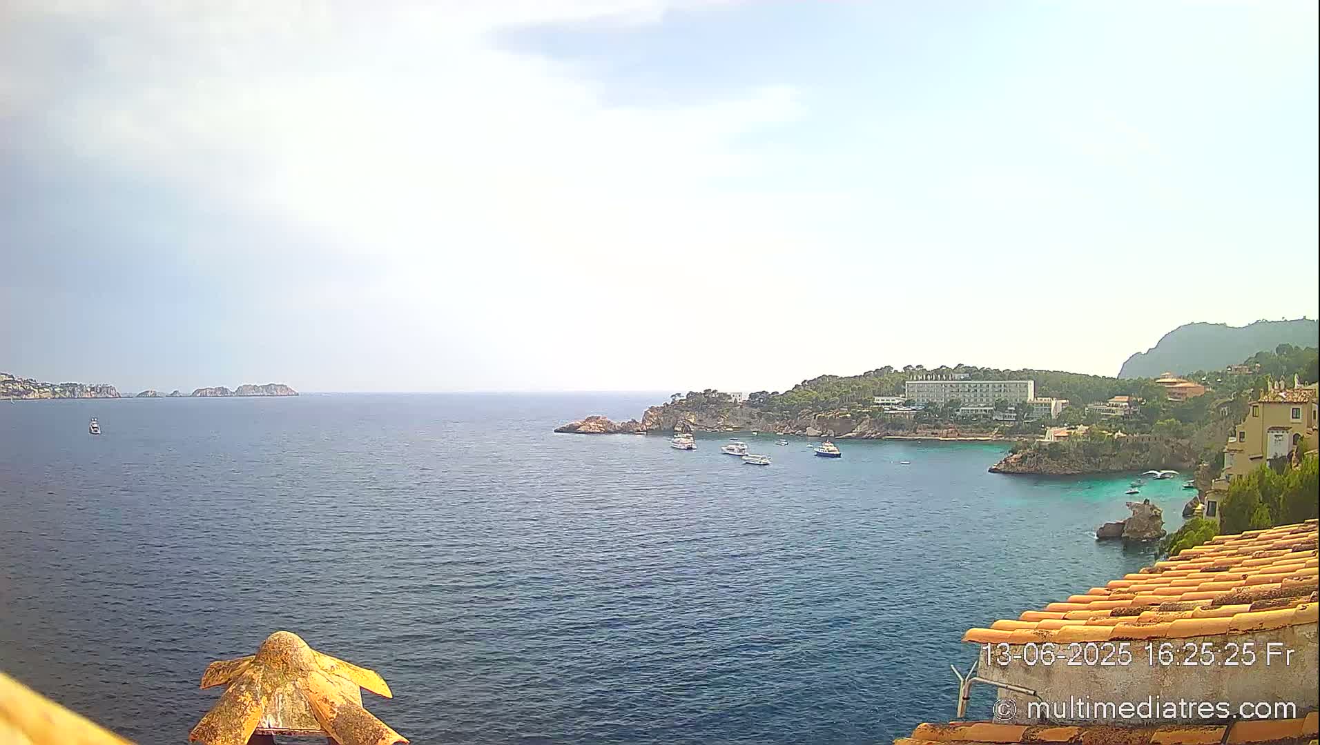 A calm, hazy day reveals a picturesque coastal scene with a tranquil bay featuring several boats, a rocky shoreline dotted with buildings, and distant islands under a mostly clear sky.