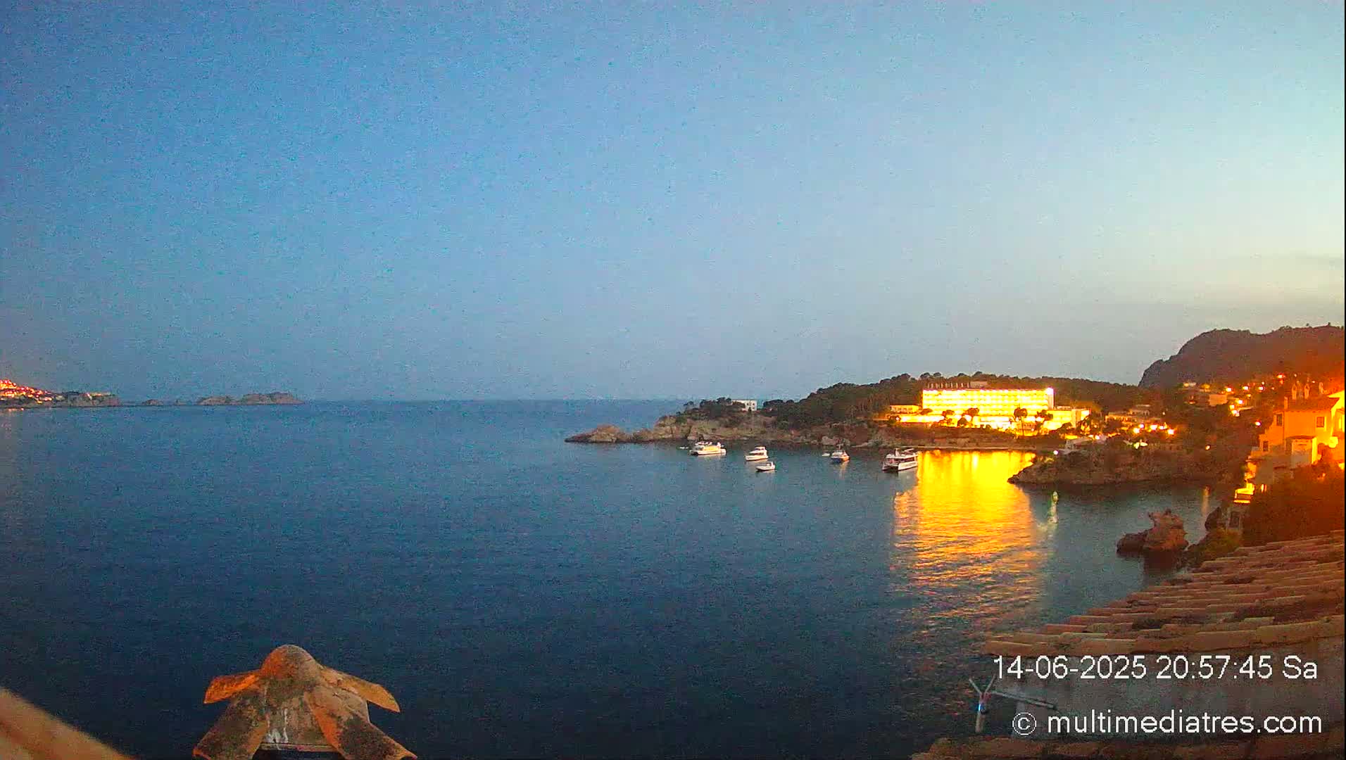 A tranquil, sunny coastal scene shows a calm blue sea dotted with boats, and a coastline with buildings and vegetation under a clear sky.