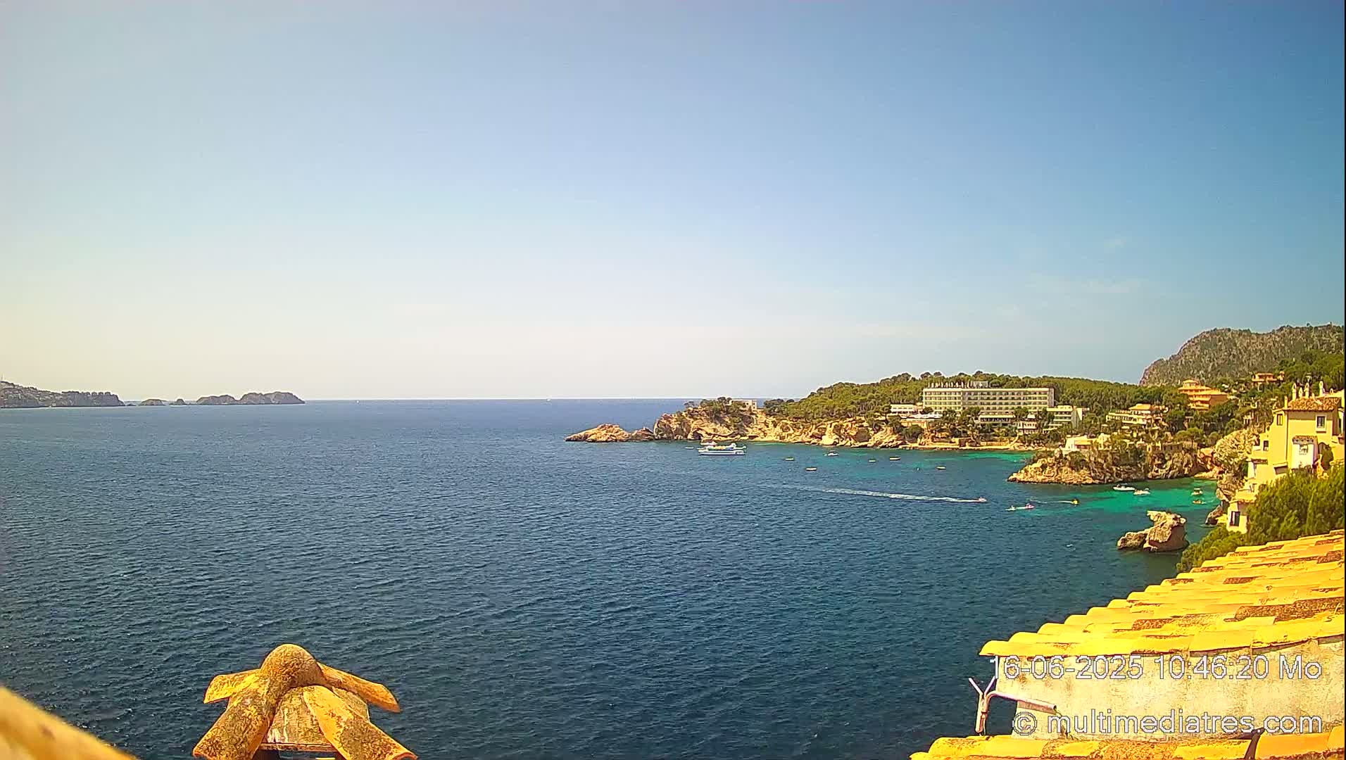 A sunny, clear day reveals a coastal view of a tranquil blue sea, dotted with boats near a rocky shoreline and buildings nestled into the hillside.