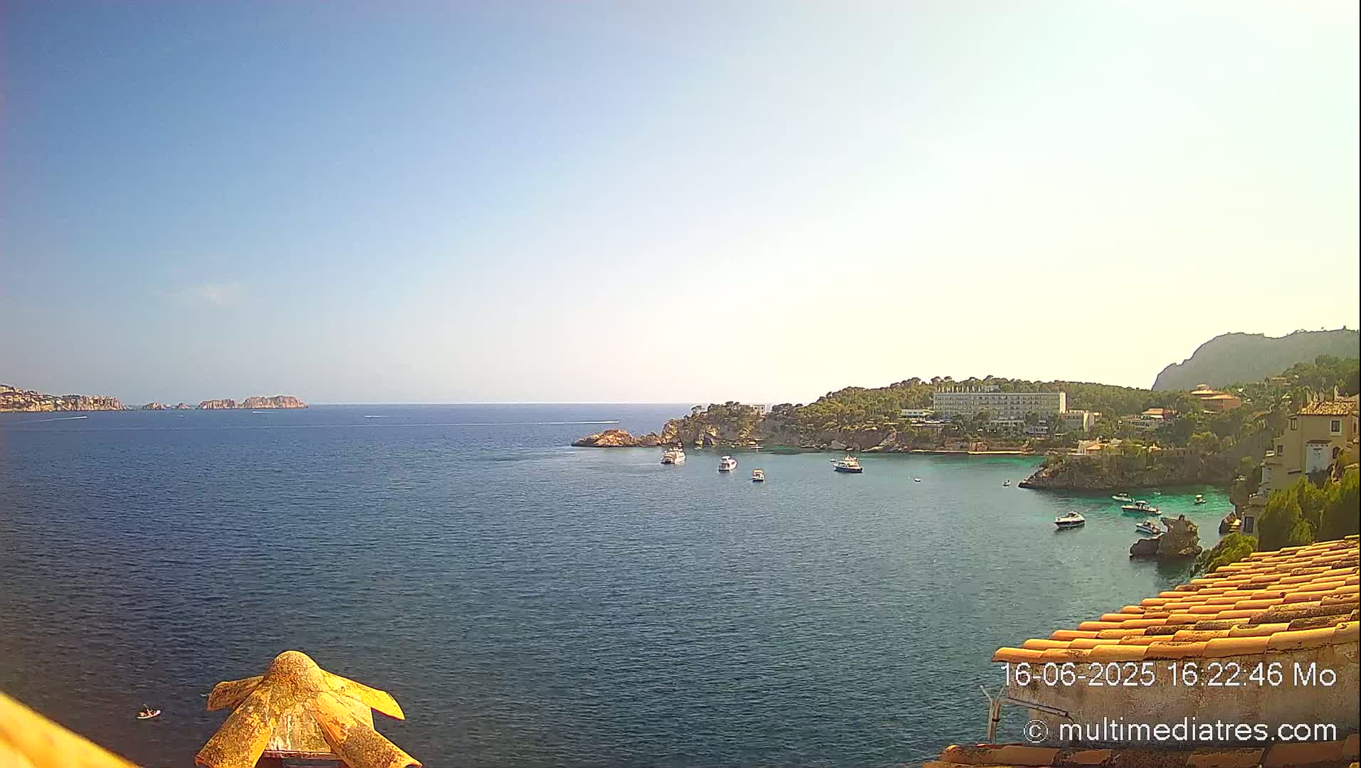 A calm, sunny day reveals a picturesque coastal scene with several boats anchored in a sheltered bay, near rocky cliffs and buildings.