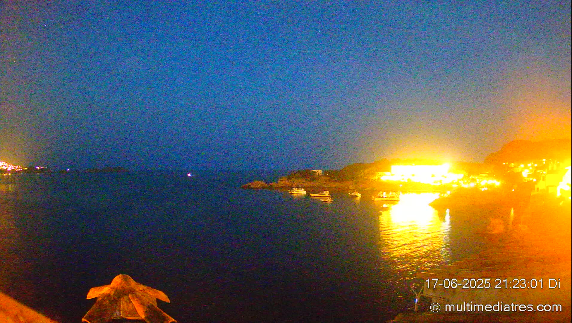 A nighttime view of a calm body of water with brightly lit buildings and boats on the shore under a dark blue sky.