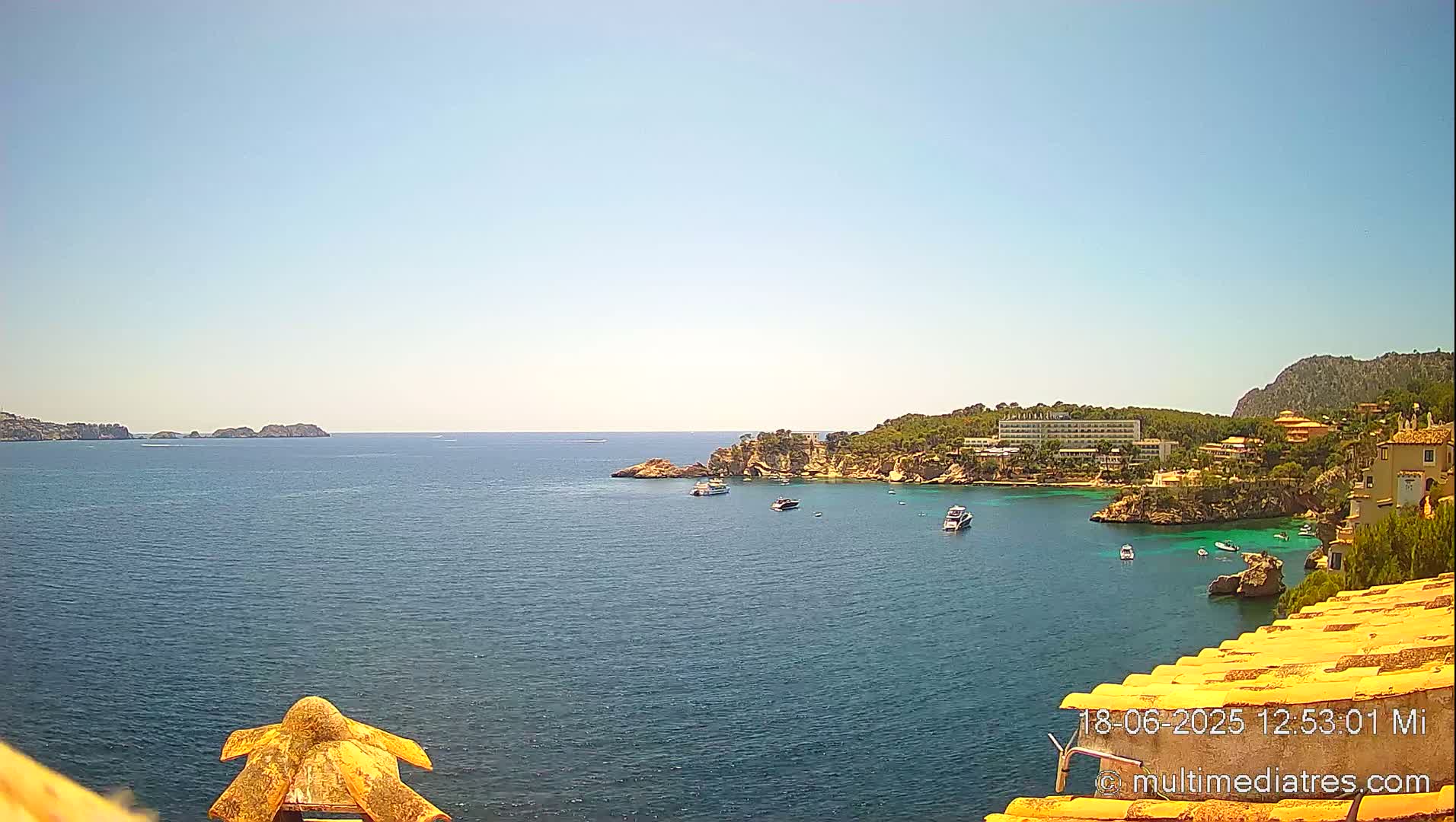 A sunny, clear day overlooking a calm bay with several boats, a hotel, and buildings nestled on a rocky coastline.