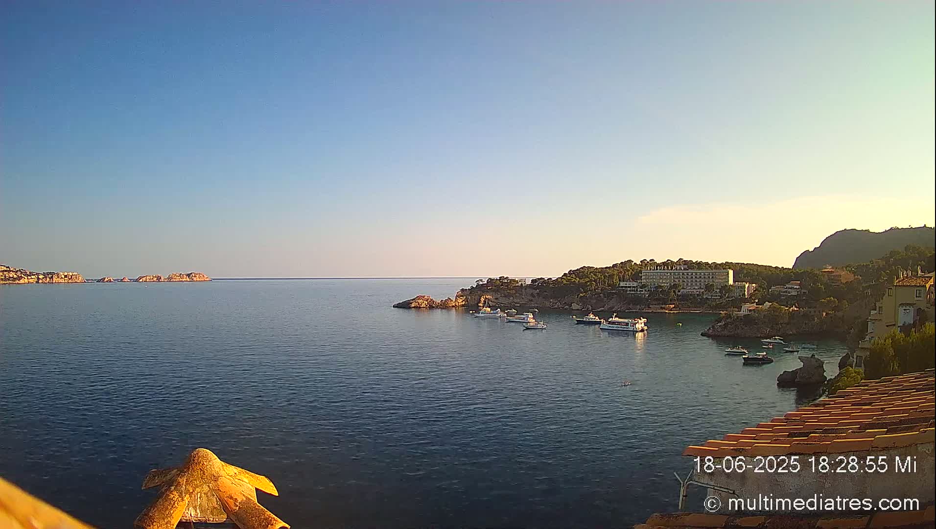 A calm, clear day reveals a coastal scene with several boats in a bay, nestled between rocky shores and a distant island under a clear blue sky.