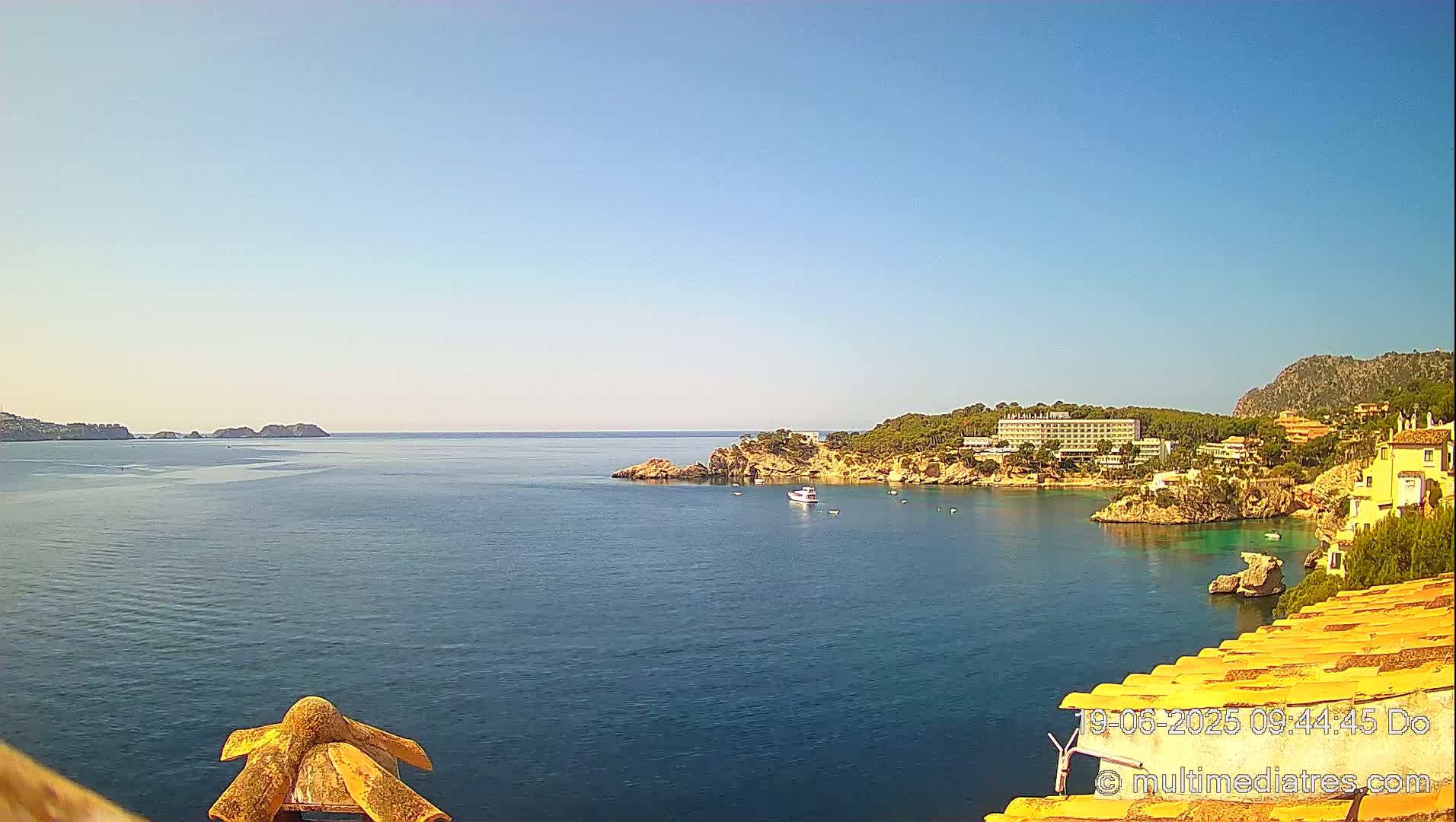 A calm, blue ocean bay is seen from a high vantage point, with a hotel and other buildings nestled along the rocky shoreline under a clear sky.
