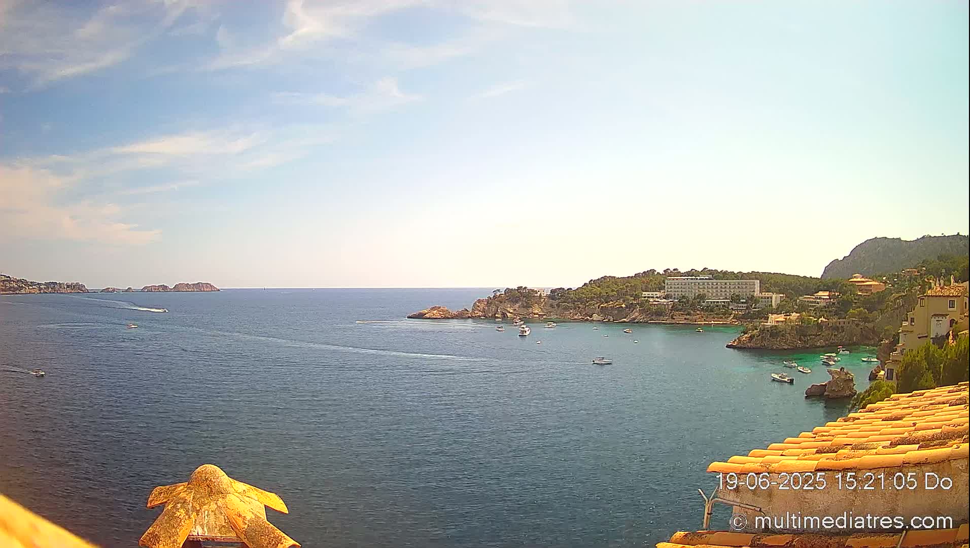 A scenic overlook of a calm, blue bay with several boats, a hotel, and rocky coastlines under a mostly sunny sky.