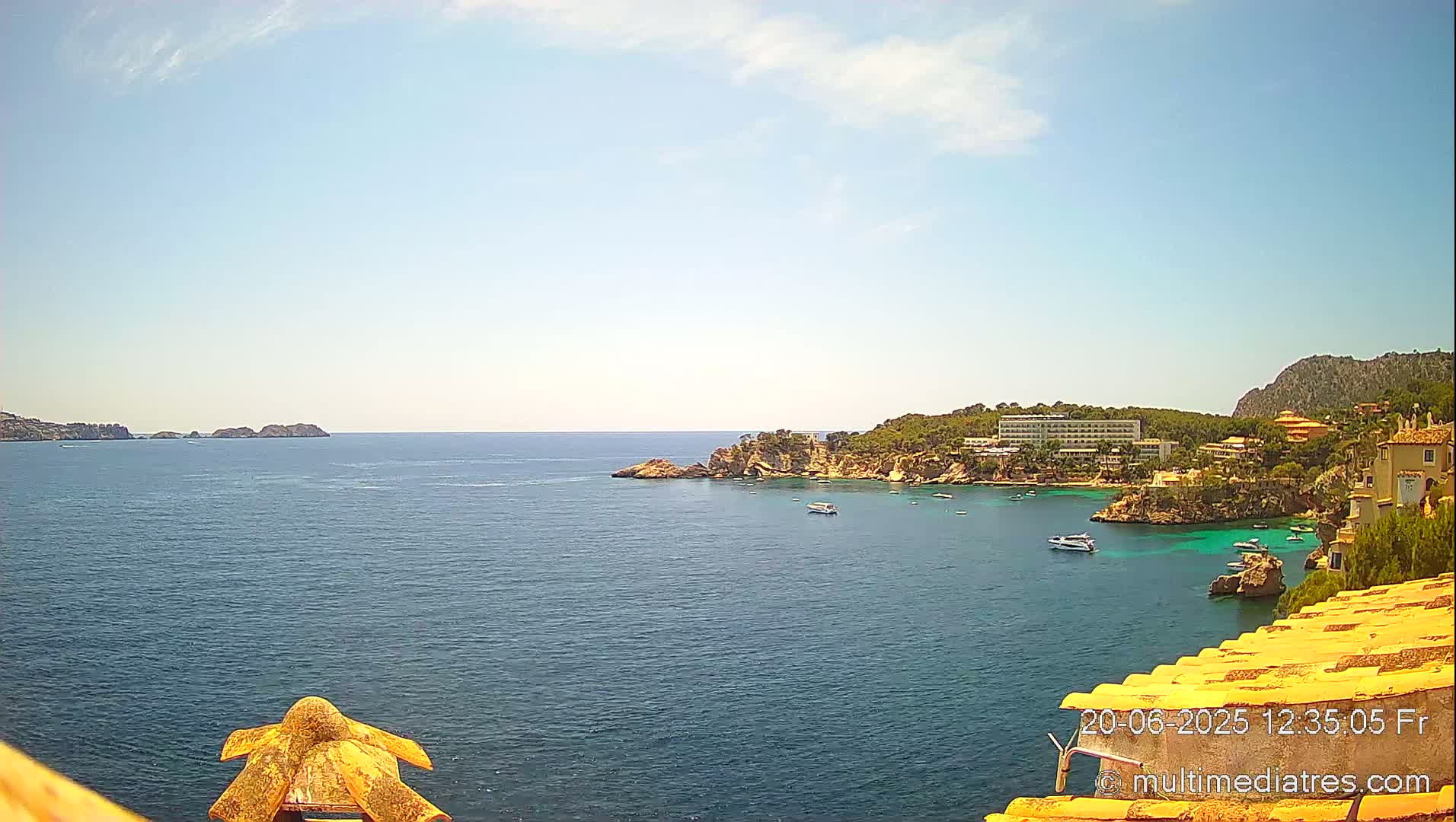 A sunny day reveals a picturesque coastal scene with calm, blue water, a rocky shoreline dotted with buildings and vegetation, and several small boats.