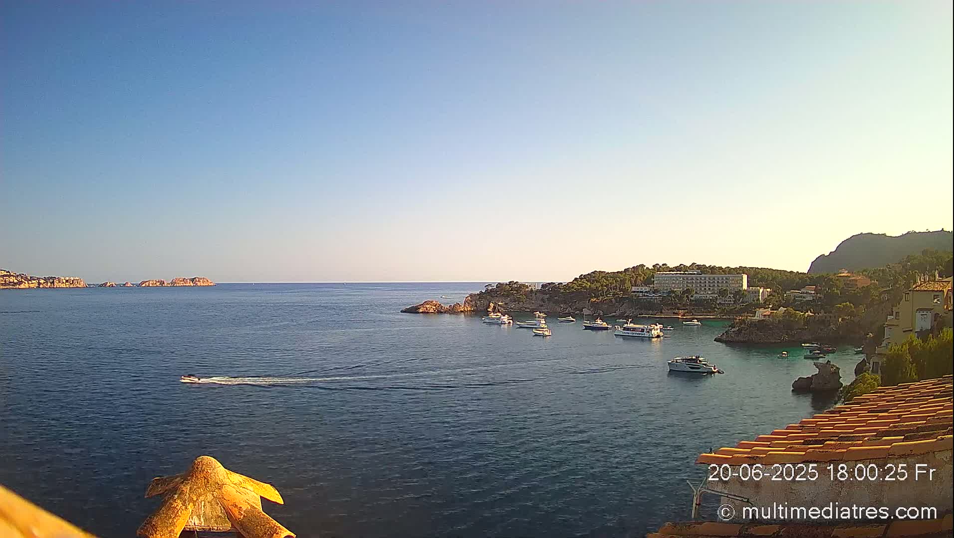 A calm, clear day reveals a picturesque coastal scene with numerous boats in a bay near a rocky shore and buildings, under a clear blue sky.