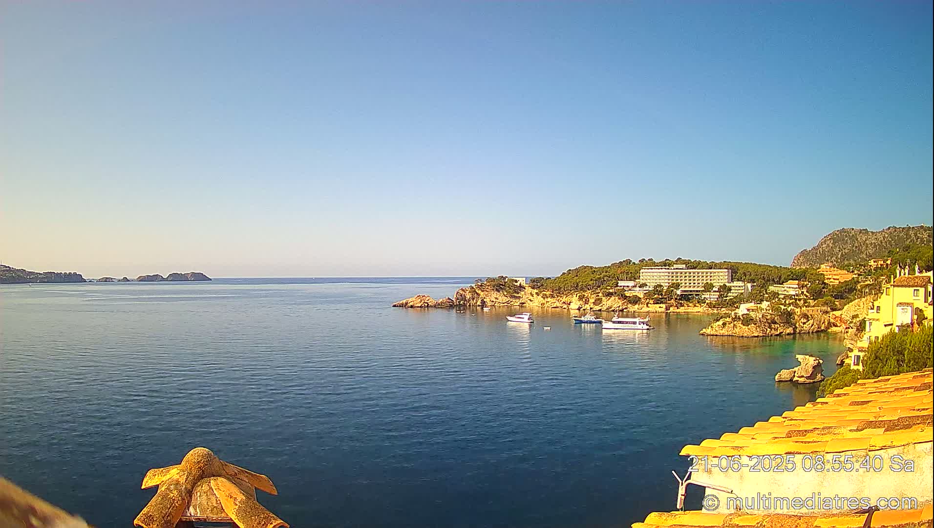 A calm, clear blue sea with a rocky coastline featuring buildings and boats under a bright, sunny sky.