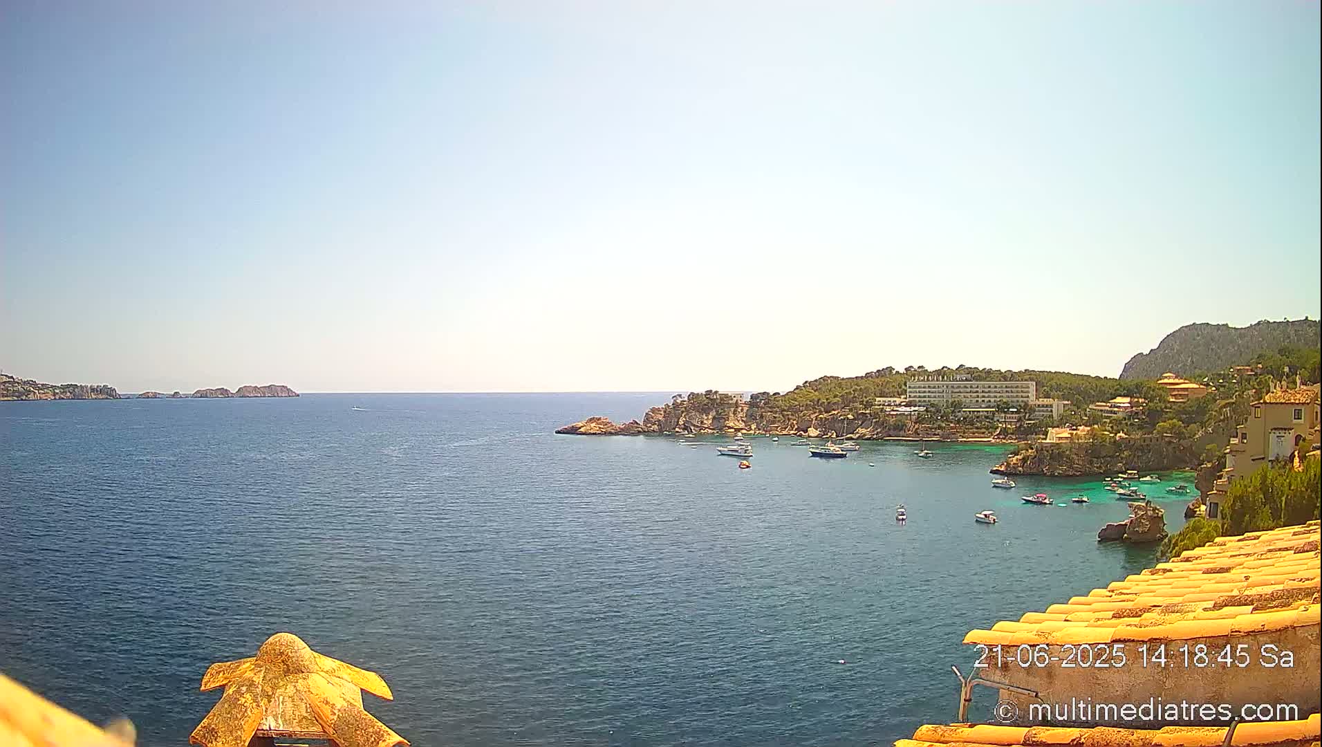 A sunny, clear day shows a calm sea with several boats anchored near a rocky coastline featuring buildings and vegetation.