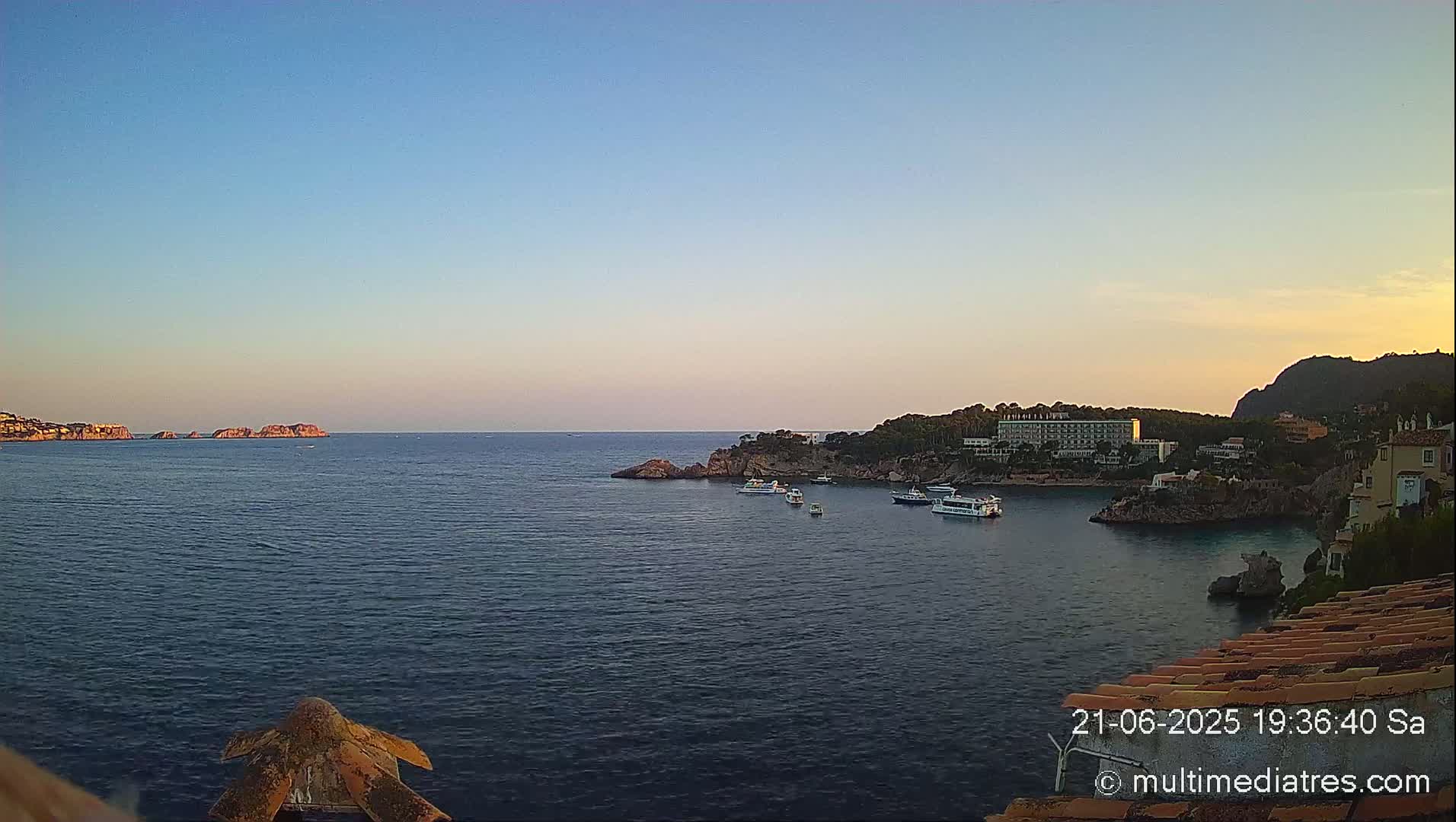 A calm, twilight sea scene shows a hotel and buildings nestled into a rocky cove, with several boats anchored in the bay and small islands visible in the distance.