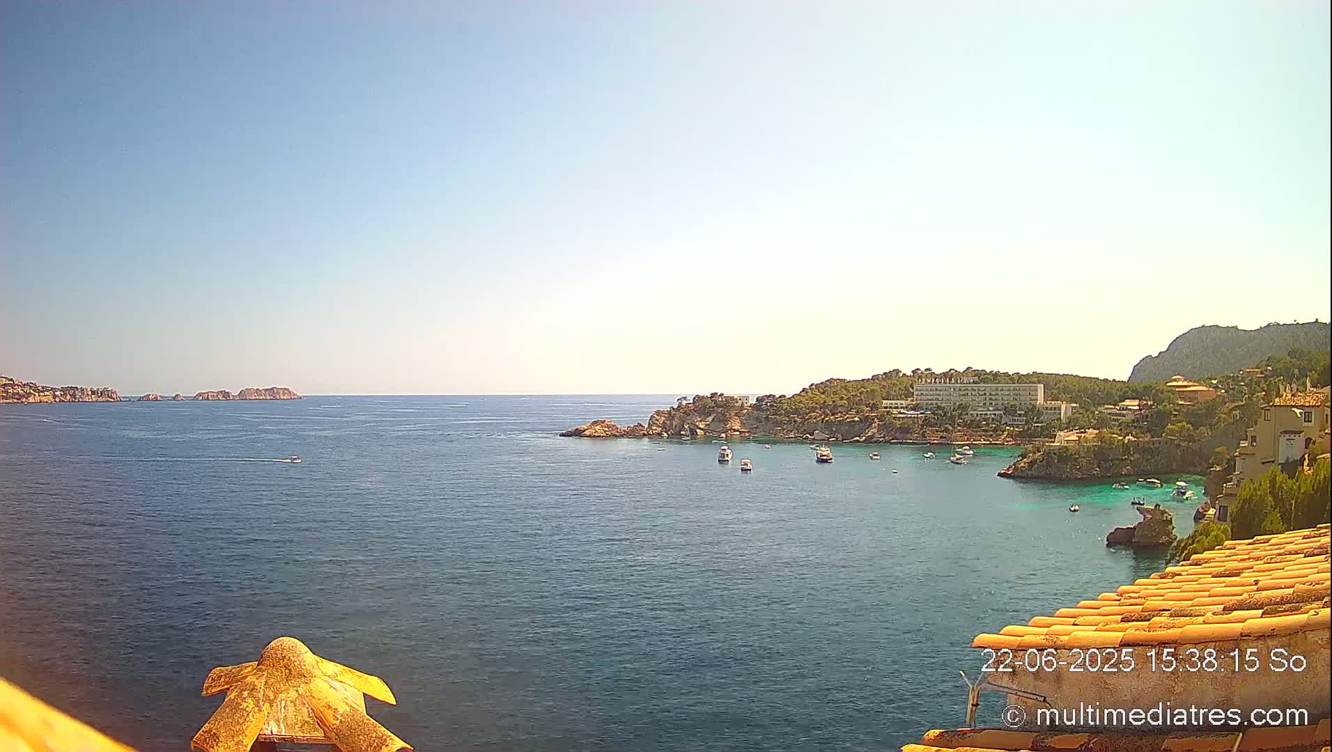 A clear, sunny day reveals a tranquil bay with several boats anchored near a coastline dotted with buildings, all under a mostly clear sky.
