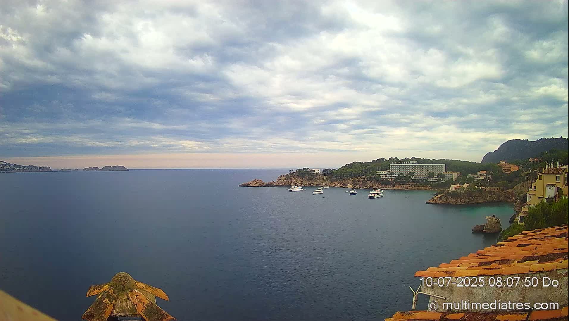 A coastal scene under a partly cloudy sky shows a calm body of water with several boats, a rocky shoreline with buildings and trees, and a distant mountain.