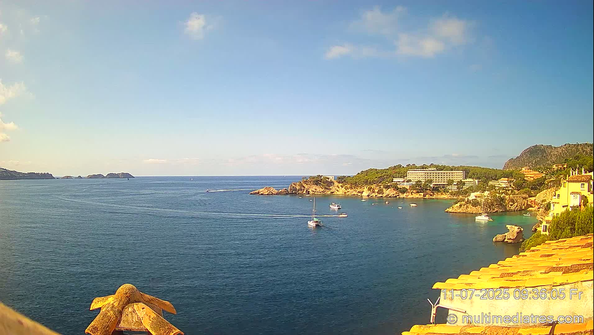 A calm, sunny day reveals a picturesque coastal scene with a tranquil bay filled with boats, nestled between a rocky shoreline and a distant mountain.