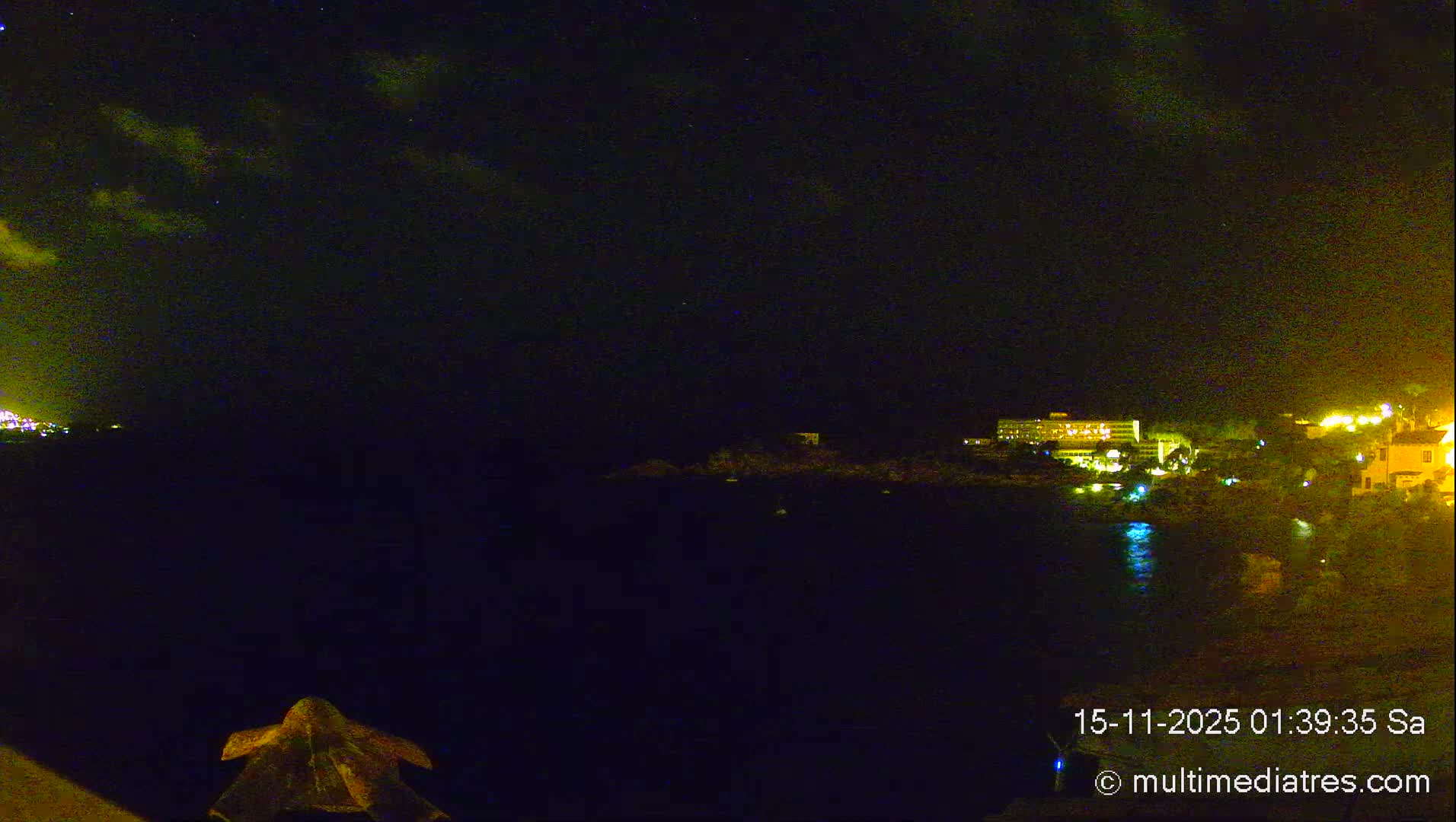 A dark night scene reveals a well-lit coastal town with buildings reflecting in calm water under a partly cloudy sky with faint stars.