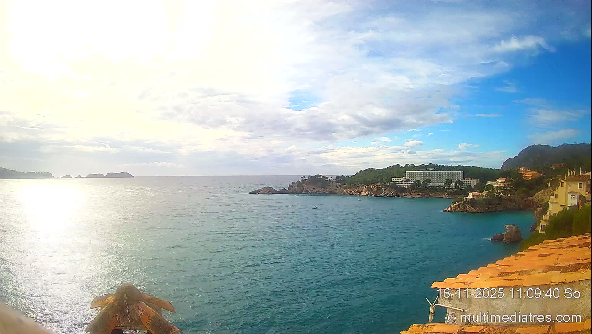 Under a bright, partly cloudy sky, a scenic turquoise bay is seen, bordered by a rocky coastline featuring hotels and coastal buildings with orange tiled roofs, lush hills, and distant landmasses, with the sun brightly reflecting on the water.