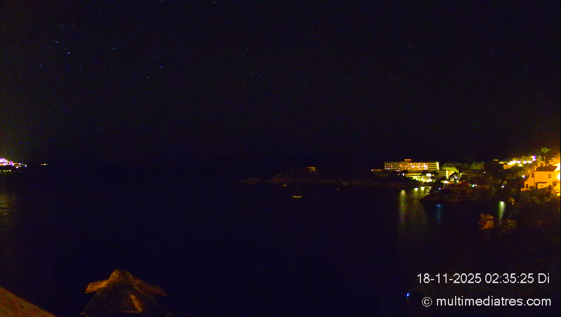 A clear, starry night sky hangs over a dark bay reflecting the lights of illuminated coastal buildings, with a few small boats visible on the calm water.