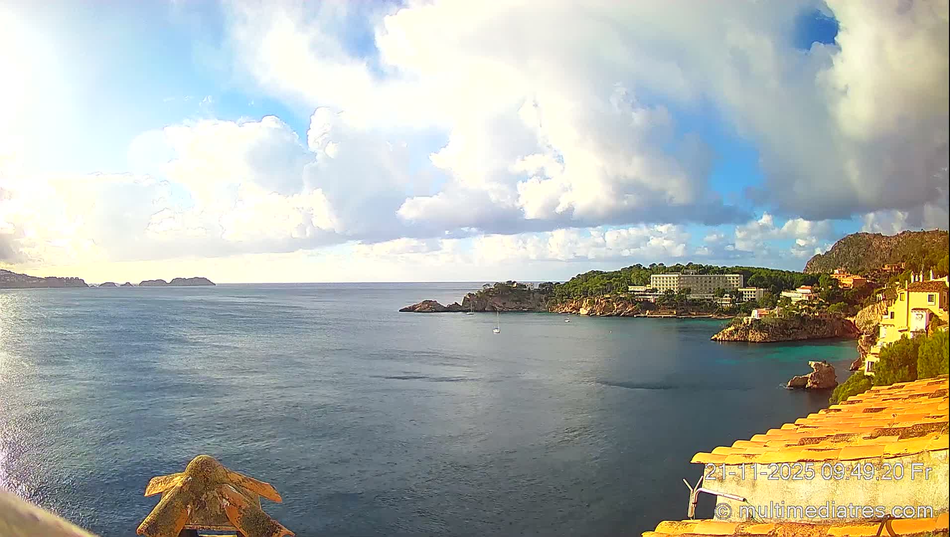 Under a bright, partly cloudy sky, a scenic coastal view features a vast blue sea with hints of turquoise in a cove, dotted with small boats and framed by rocky headlands, distant islands, and a town with buildings nestled into green, hilly terrain.