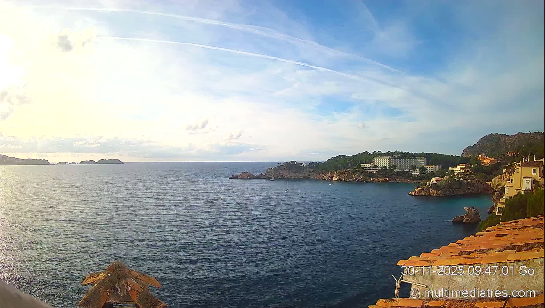 A scenic coastal view unfolds under a partly cloudy and sunny sky, showcasing a calm blue sea dotted with sailboats, a rocky coastline adorned with lush trees and buildings, and a distant mountainous backdrop.