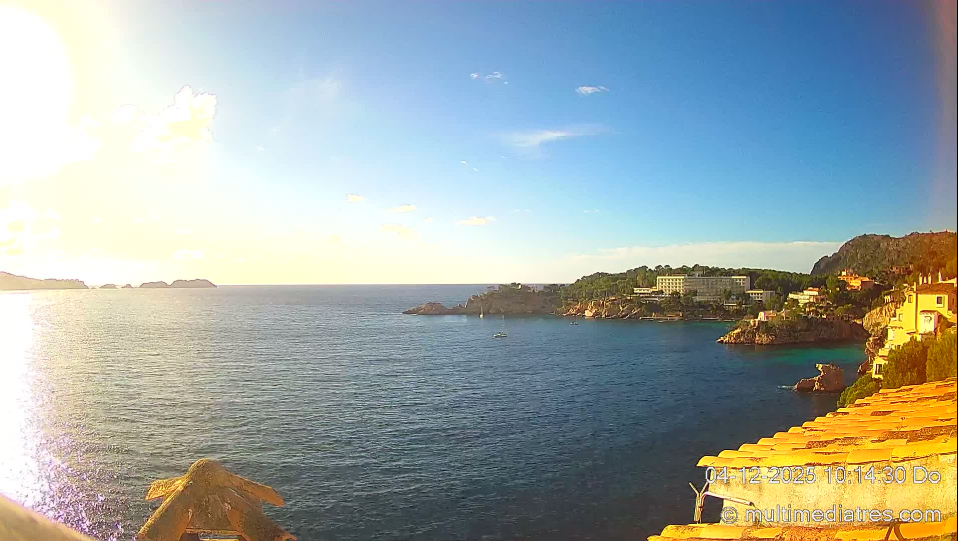 Under a clear, sunny sky, a wide expanse of deep blue-green sea stretches out, featuring rocky coves with scattered boats, a coastline adorned with buildings and lush green hills, and distant landmasses, with an orange-tiled structure visible in the foreground.