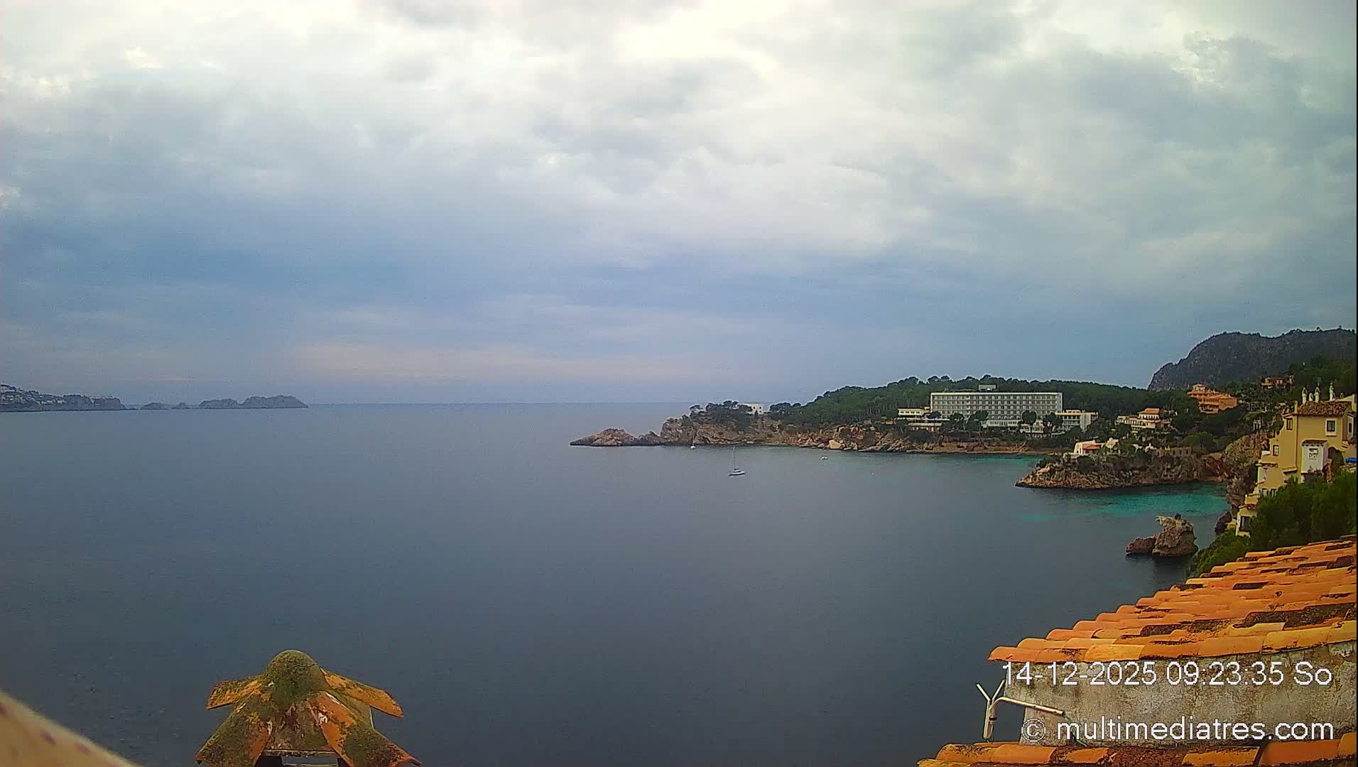 Under a clear, sunny sky, a wide expanse of deep blue-green sea stretches out, featuring rocky coves with scattered boats, a coastline adorned with buildings and lush green hills, and distant landmasses, with an orange-tiled structure visible in the foreground.