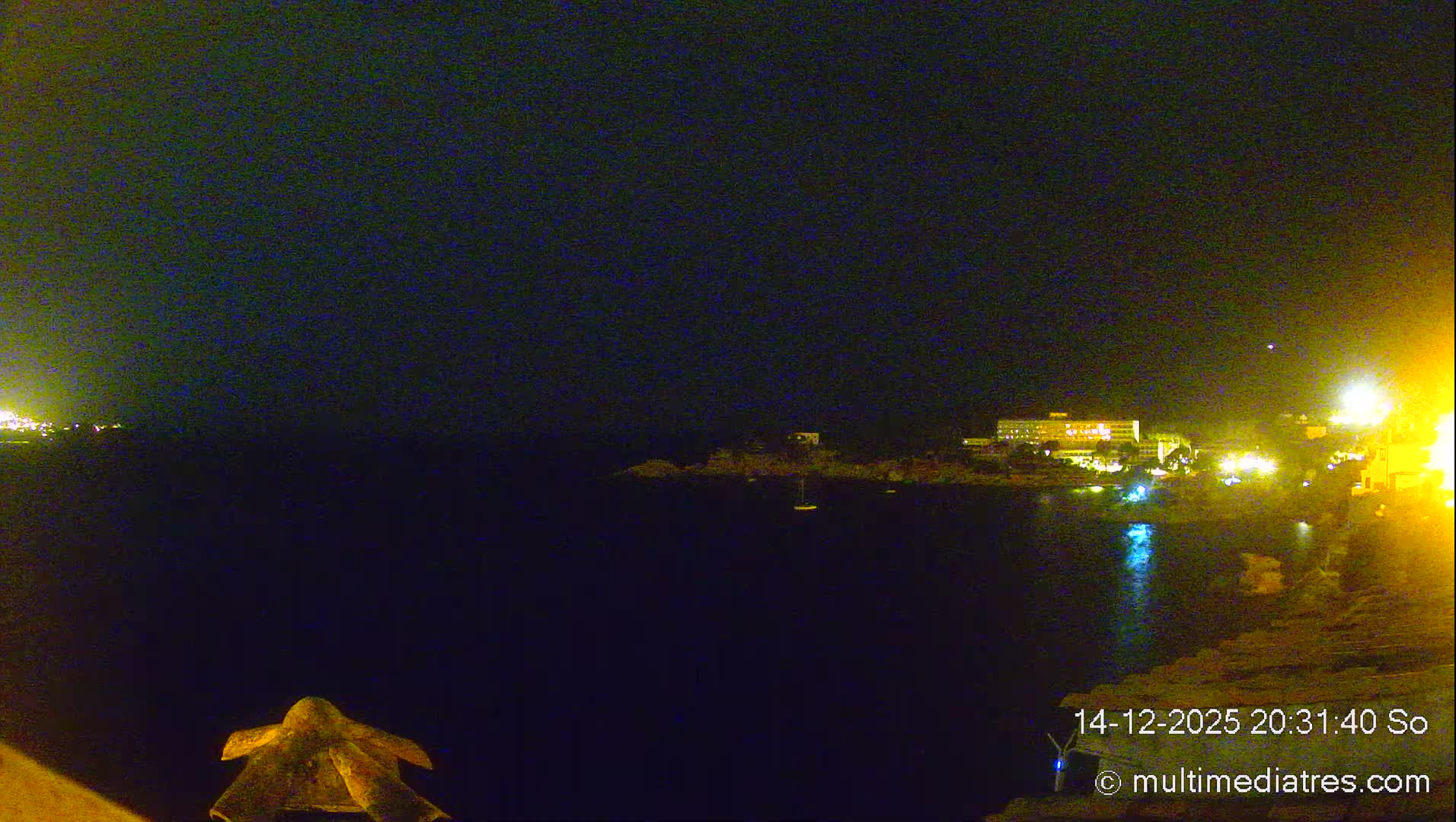 Under a clear, sunny sky, a wide expanse of deep blue-green sea stretches out, featuring rocky coves with scattered boats, a coastline adorned with buildings and lush green hills, and distant landmasses, with an orange-tiled structure visible in the foreground.