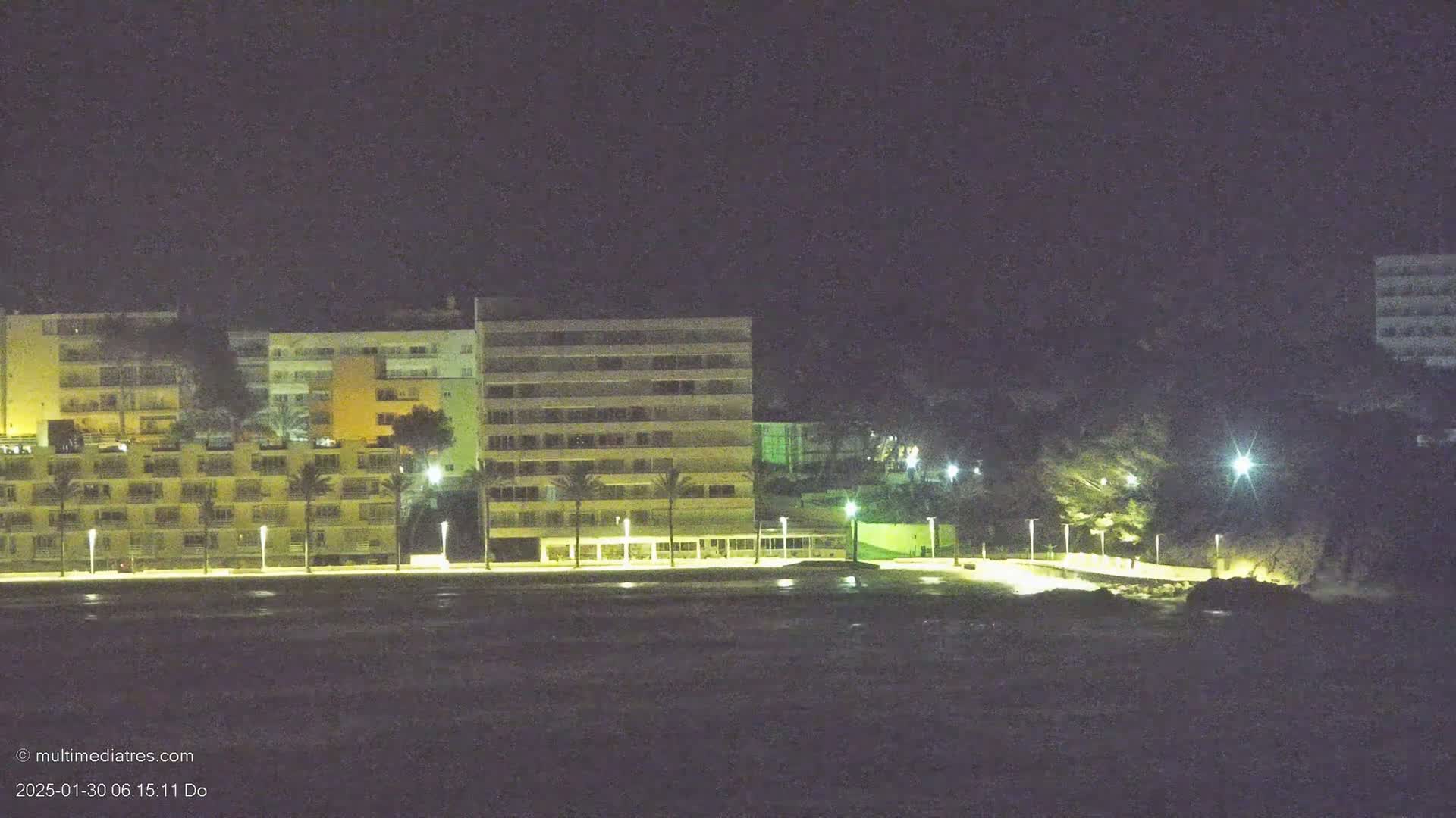At night, illuminated multi-story buildings line a dark waterfront, with palm trees and streetlights visible along a paved walkway.