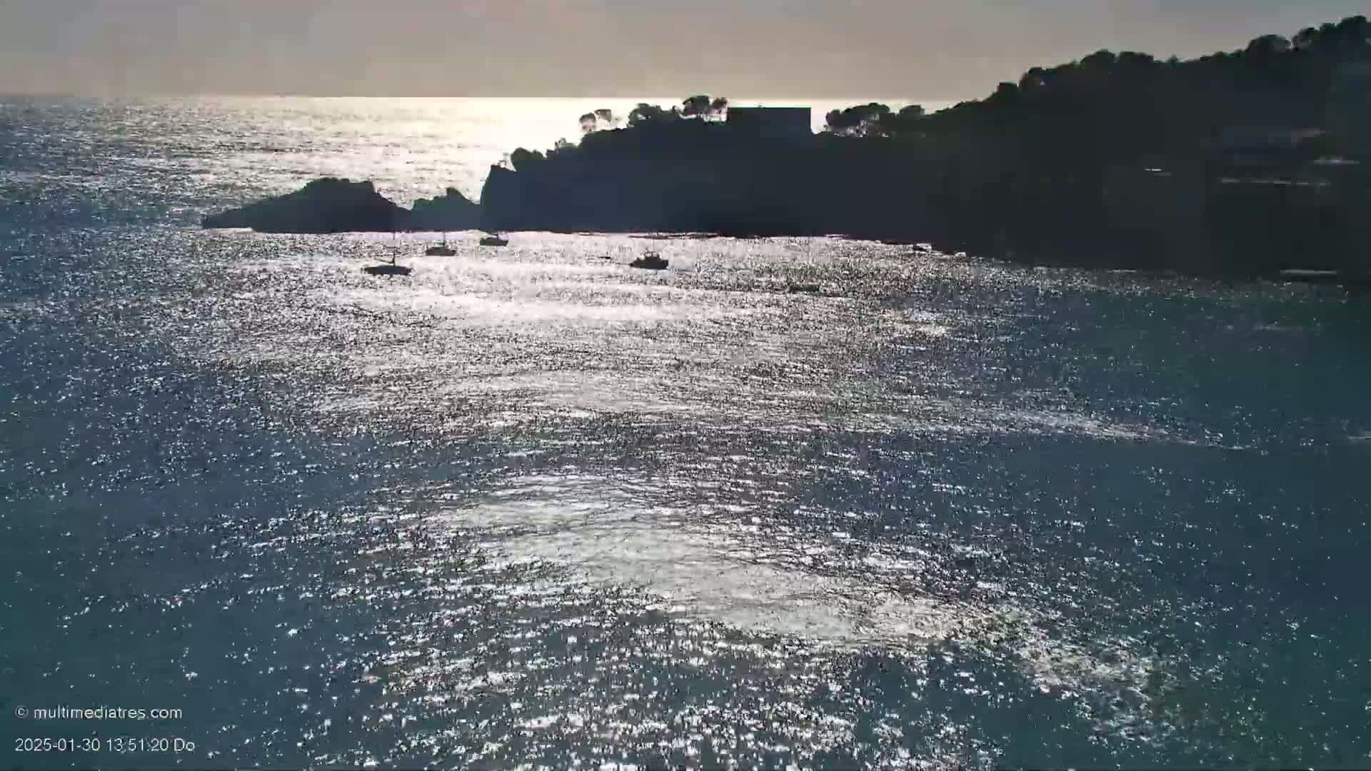 Several small boats are visible on a sparkling sea, near a dark, rocky shoreline with some trees and buildings silhouetted against a bright, but somewhat overcast sky.