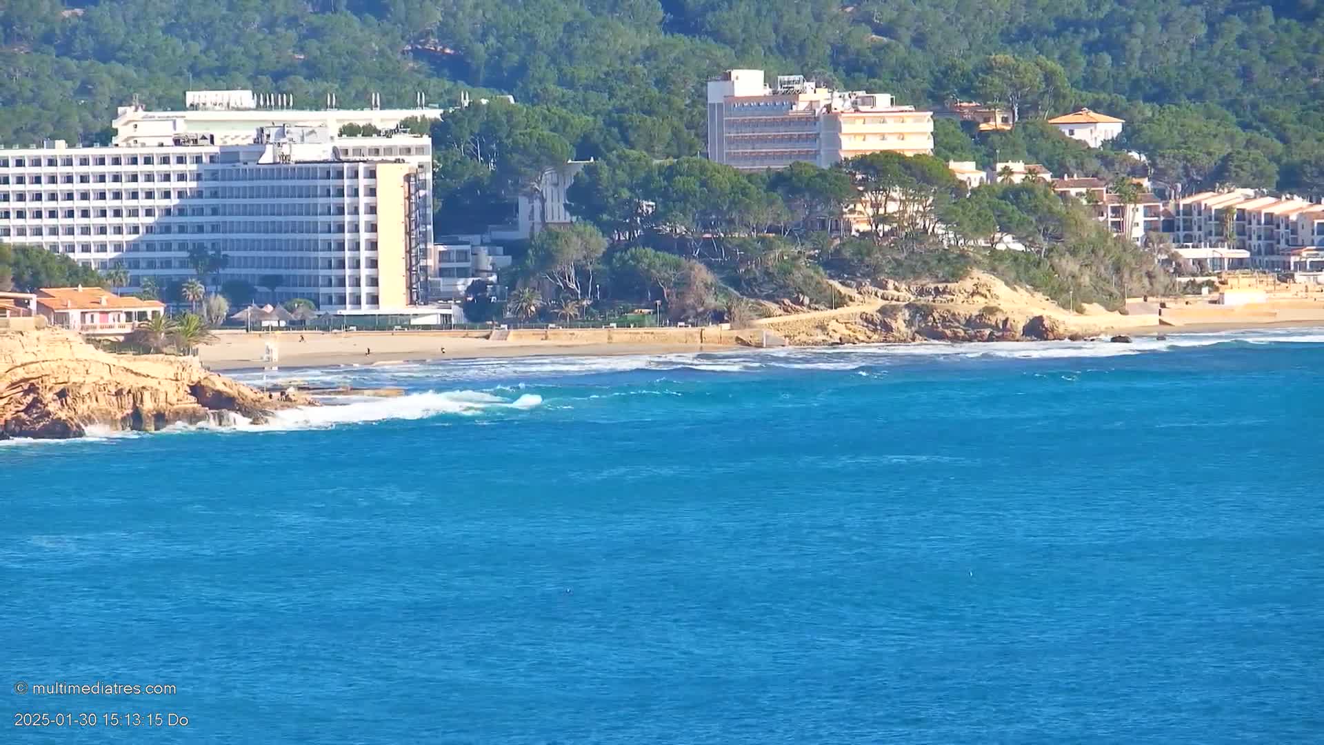 A sandy beach with several hotels and buildings sits on a hillside overlooking a blue ocean with small waves.
