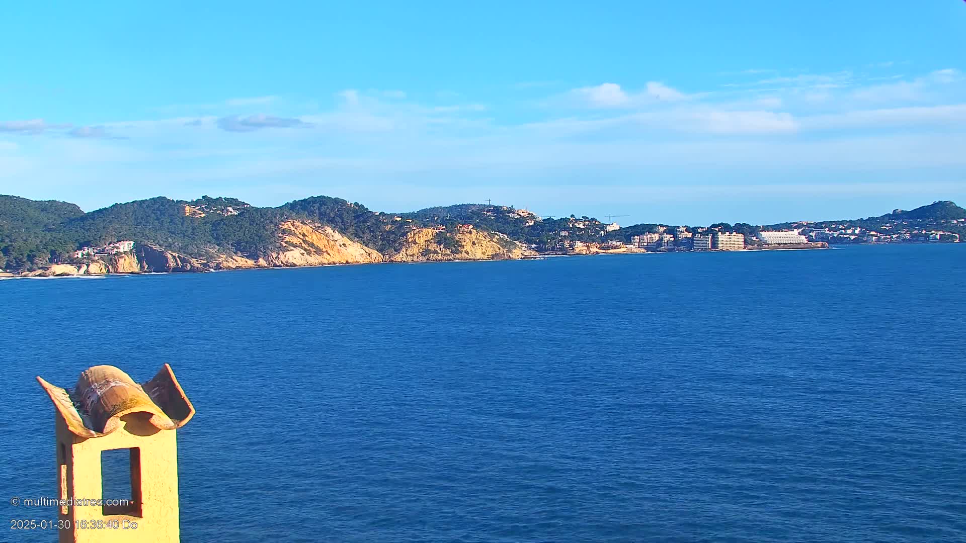 A partly cloudy blue sky overlooks a calm blue ocean, with a distant coastline featuring green hills and buildings.
