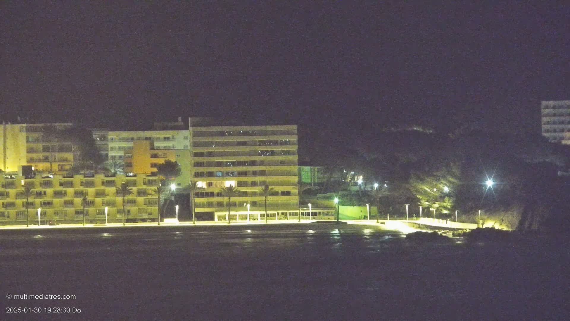 Illuminated beachfront buildings stand under a dark night sky next to a calm body of water.